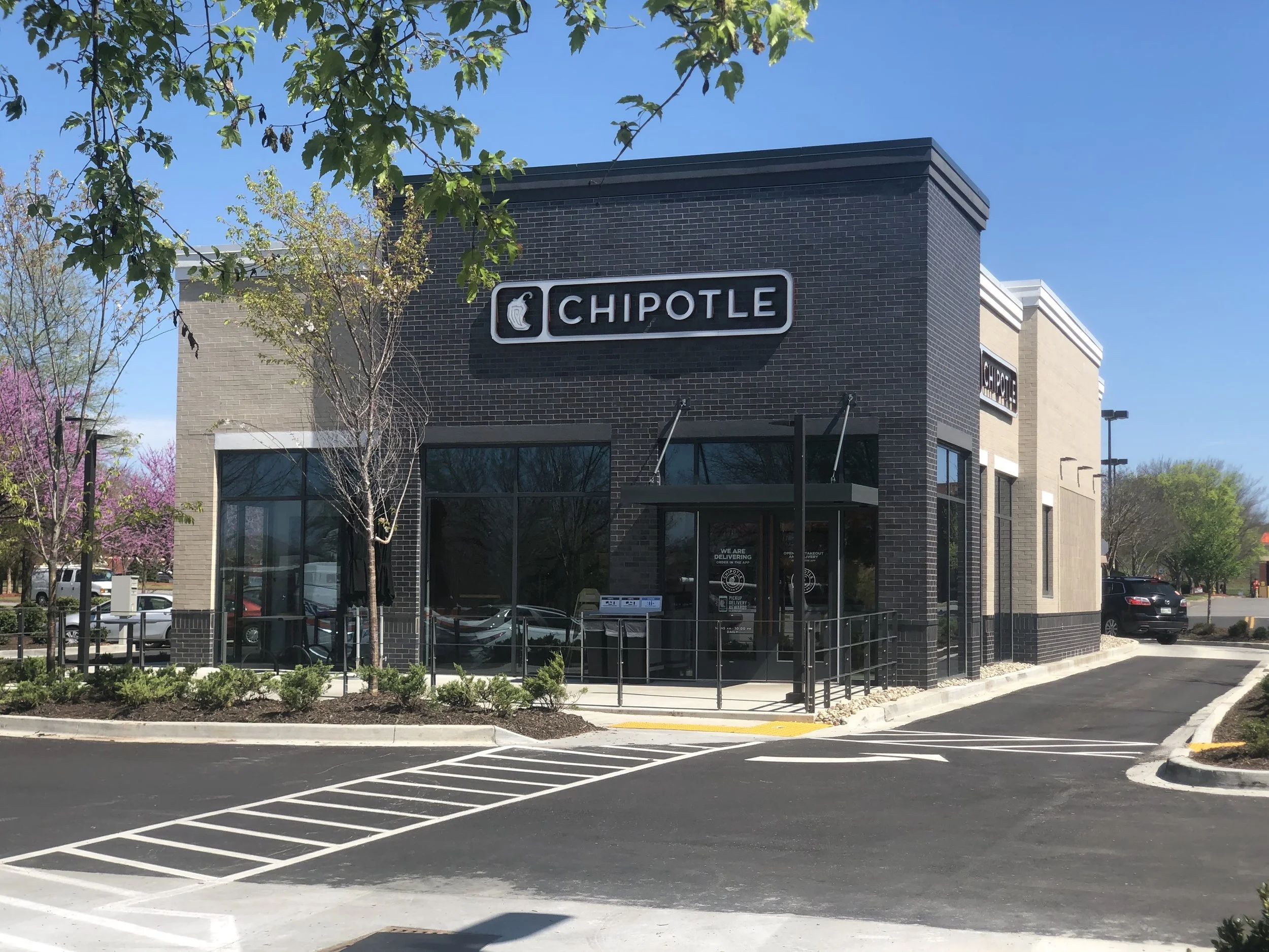 A Chipotle restaurant building with dark brick exterior, large windows, and the Chipotle logo on a sign. There is a parking lot in front with marked parking spaces and a small landscaped area with trees and bushes. The sky is clear and blue.