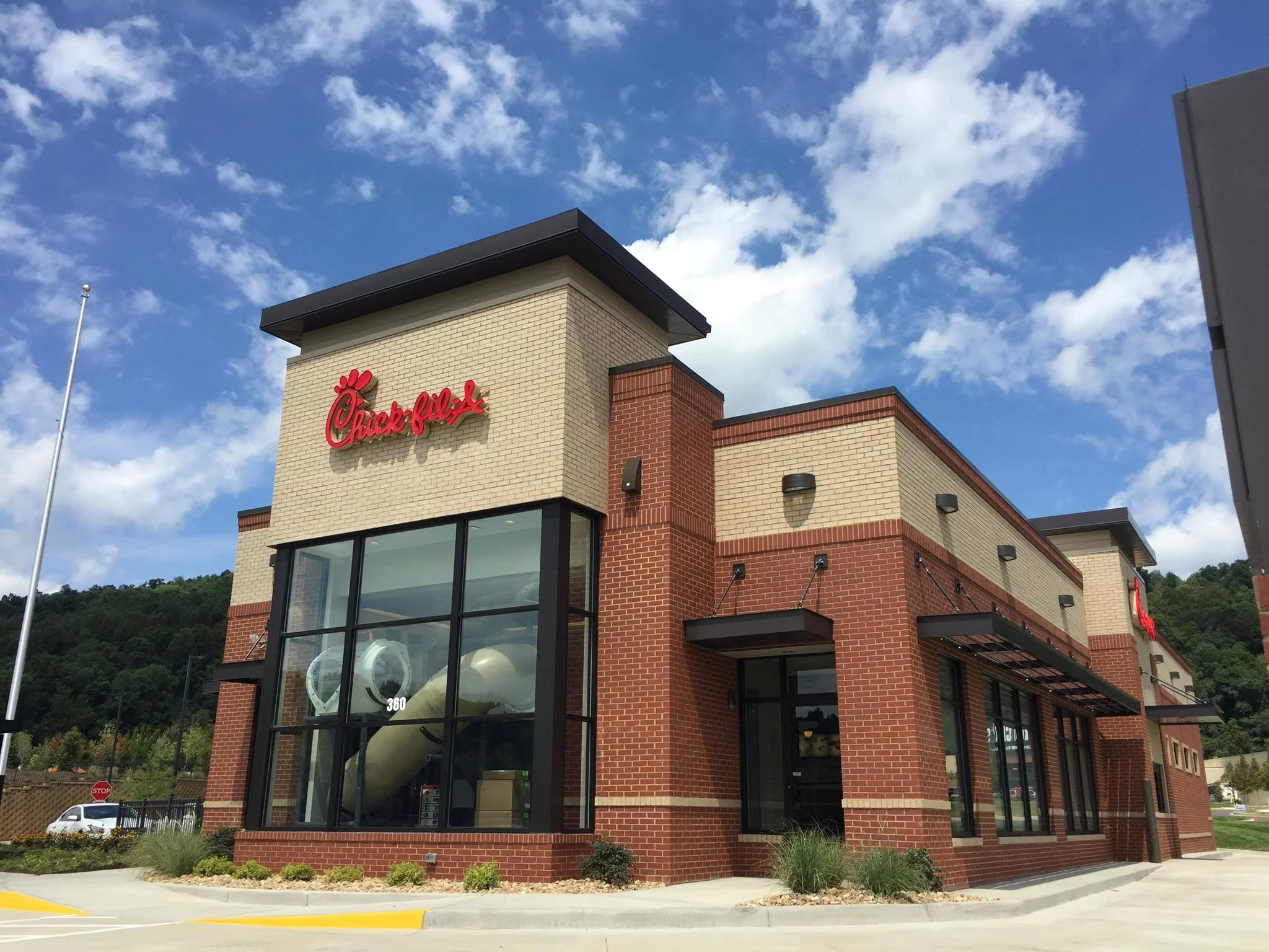 Exterior of a Chick-fil-A restaurant with a brick and glass facade under a blue sky with scattered clouds, surrounded by landscaping and parking area.