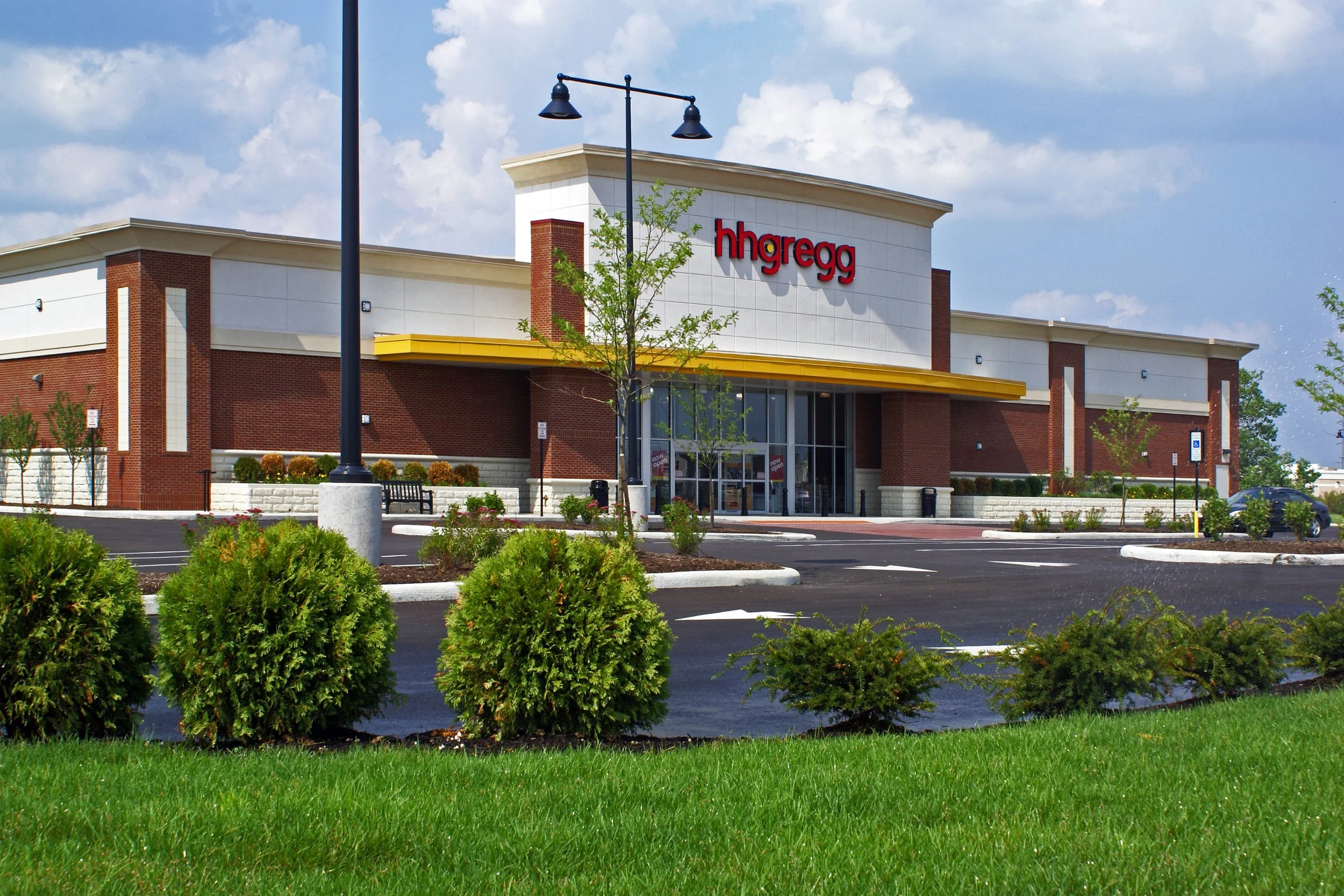 Exterior of a HomeGoods store with a red sign, large glass windows, and a yellow awning, surrounded by parking lot and landscaped bushes under a partly cloudy sky.