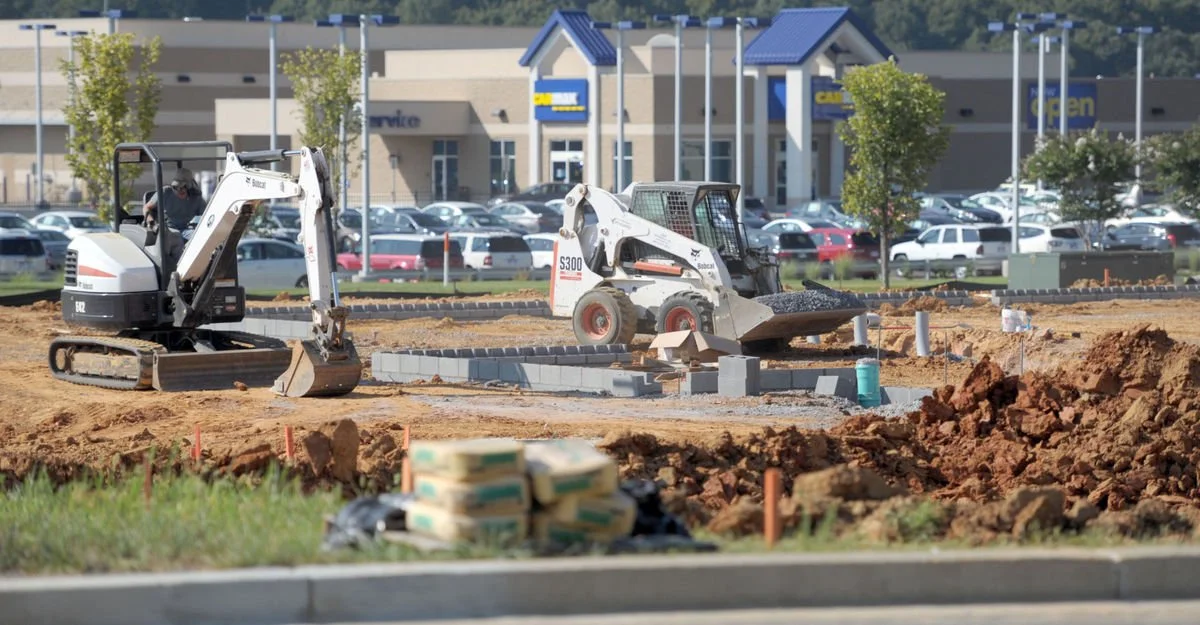 Construction site with two small excavators working on laying bricks, in front of a shopping mall parking lot with cars, trees, and a building with blue roofing.