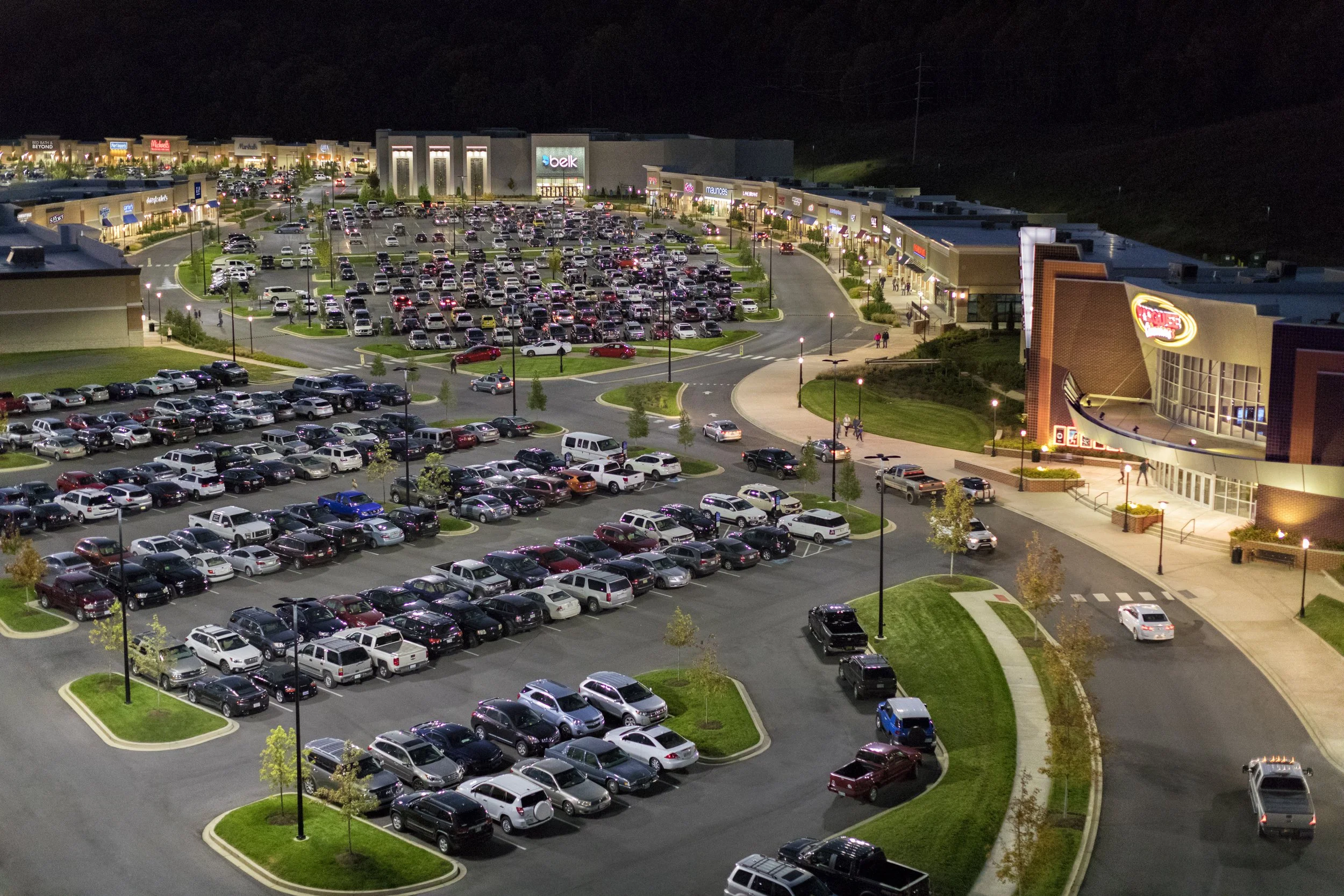 Nighttime view of a shopping mall parking lot filled with numerous cars, with illuminated storefronts and signs including Belk, Maurices, and other retail stores. A few people are walking near the entrances, with surrounding greenery and trees, and d