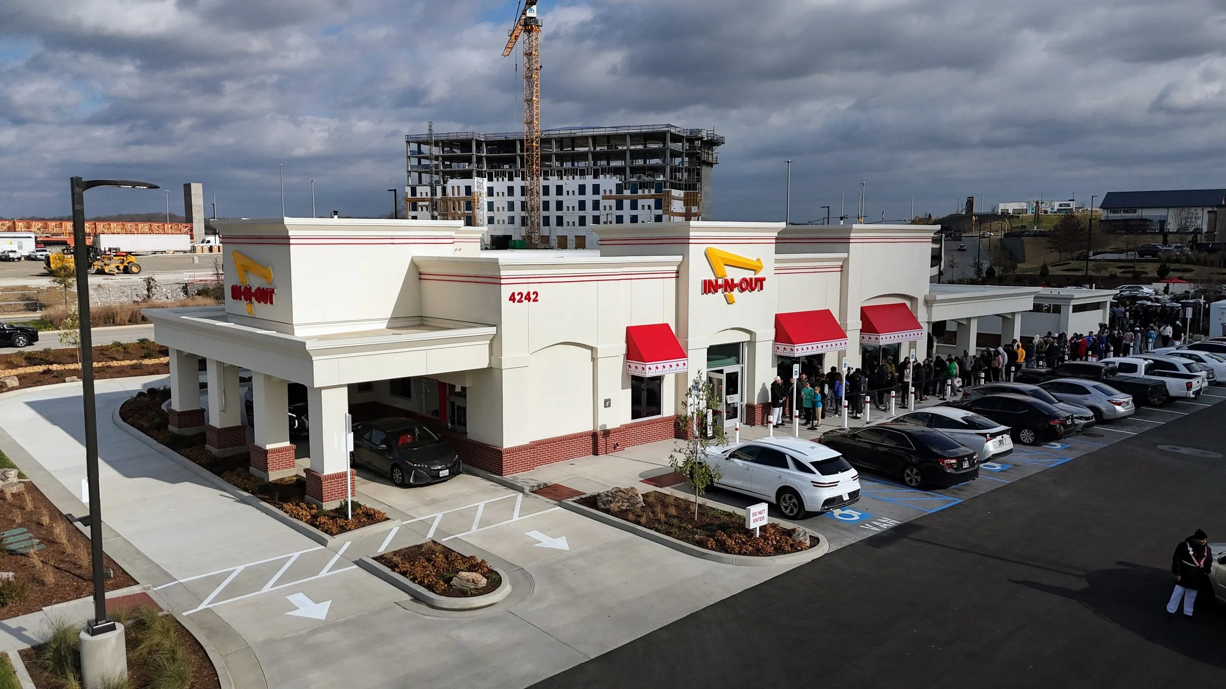 In-N-Out Burger restaurant with a long line of customers outside, parking lot with cars, and a construction site with a tall building and crane in the background.