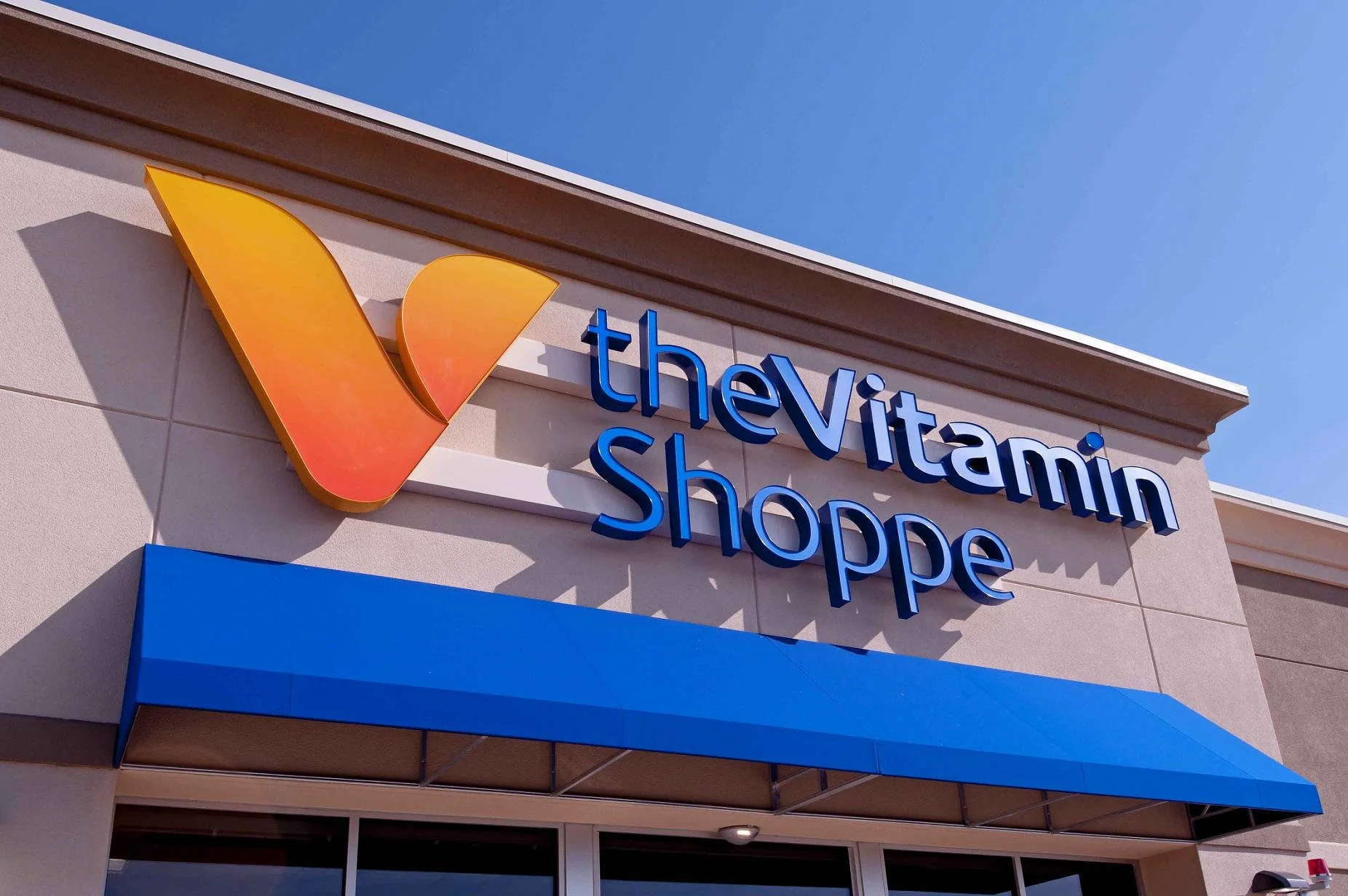 Exterior of a shopping center with a sign reading 'The Vitamin Shoppe' and a colorful vitamin logo on the building.
