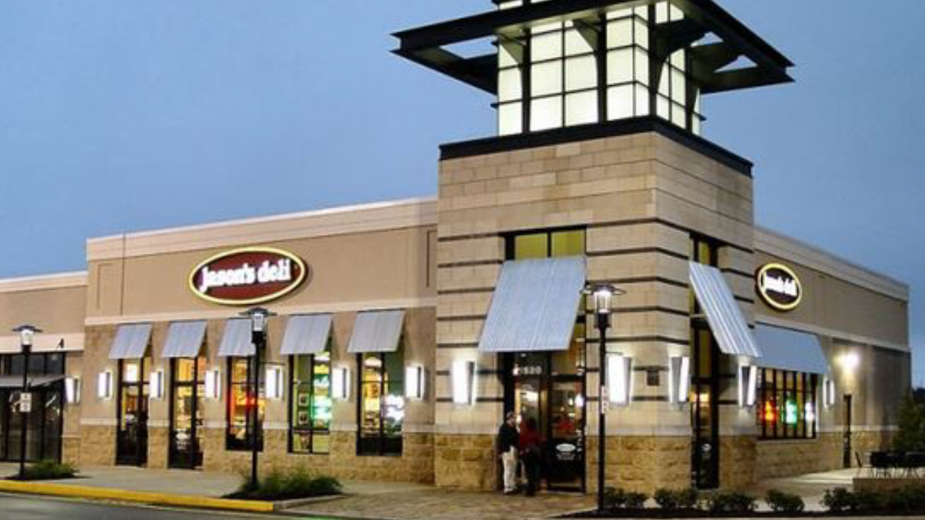 Photo of a Jack's Deli restaurant exterior at dusk, featuring a beige building with large windows, blue awnings, and a prominent tower structure with a illuminated lamp on top.