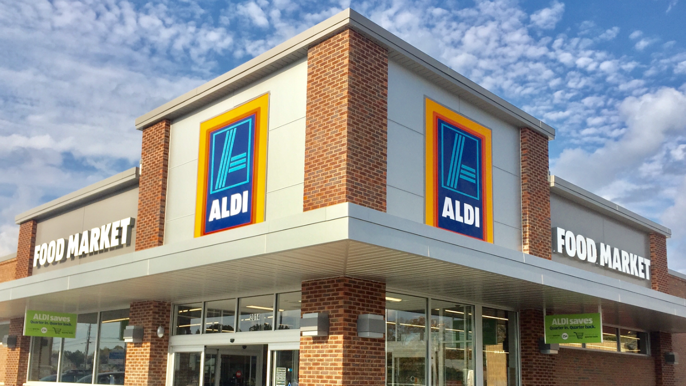Exterior view of an ALDI grocery store with brick walls and large signboards displaying ALDI logo and 'Food Market' signs, under a partly cloudy sky.