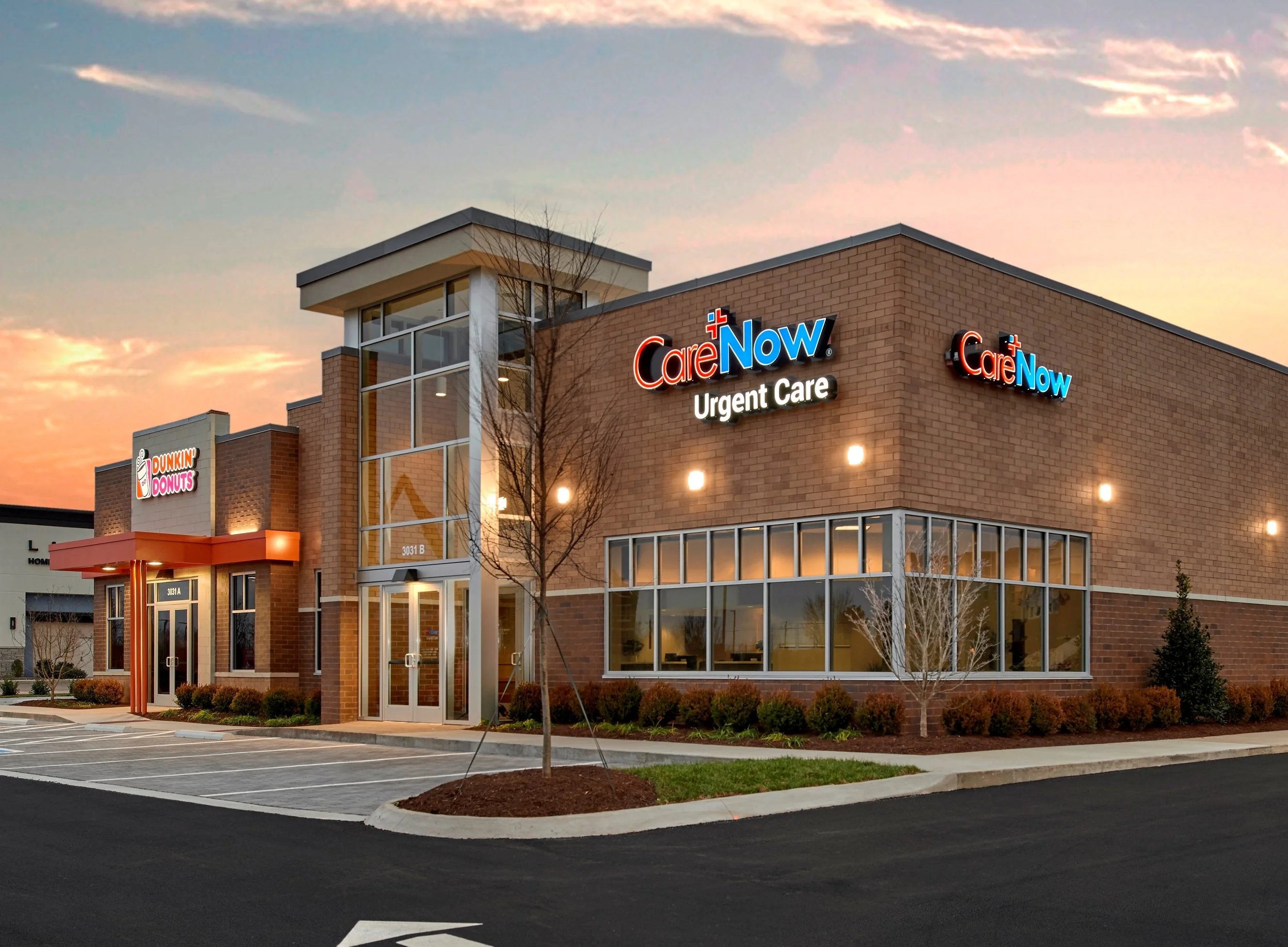 Exterior view of a Care Now urgent care center at sunset, with parking lot in foreground and trees along the building.