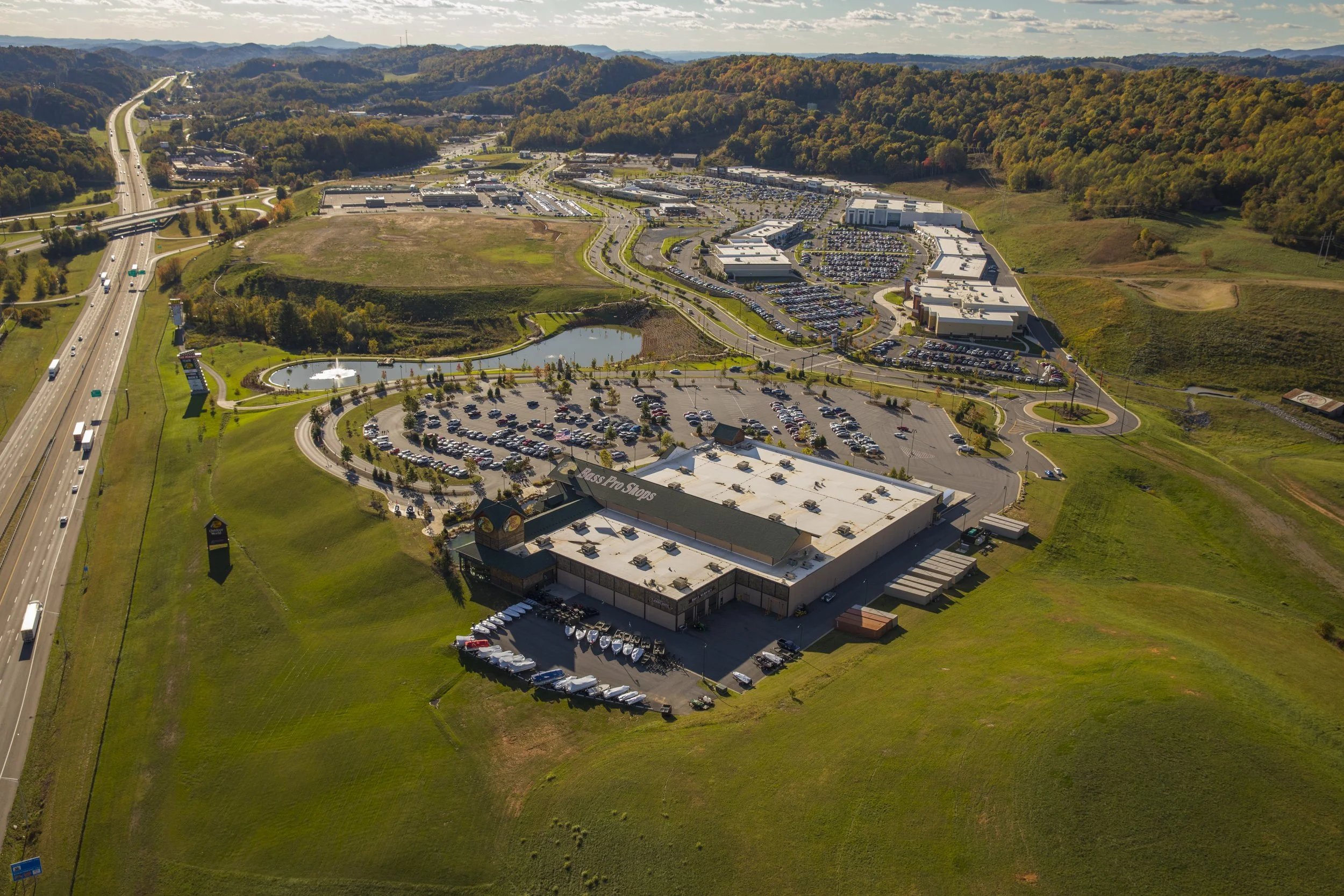 Aerial view of a shopping center surrounded by parking lots, with a pond and a fountain in front. Nearby are green hills and a highway with vehicles.