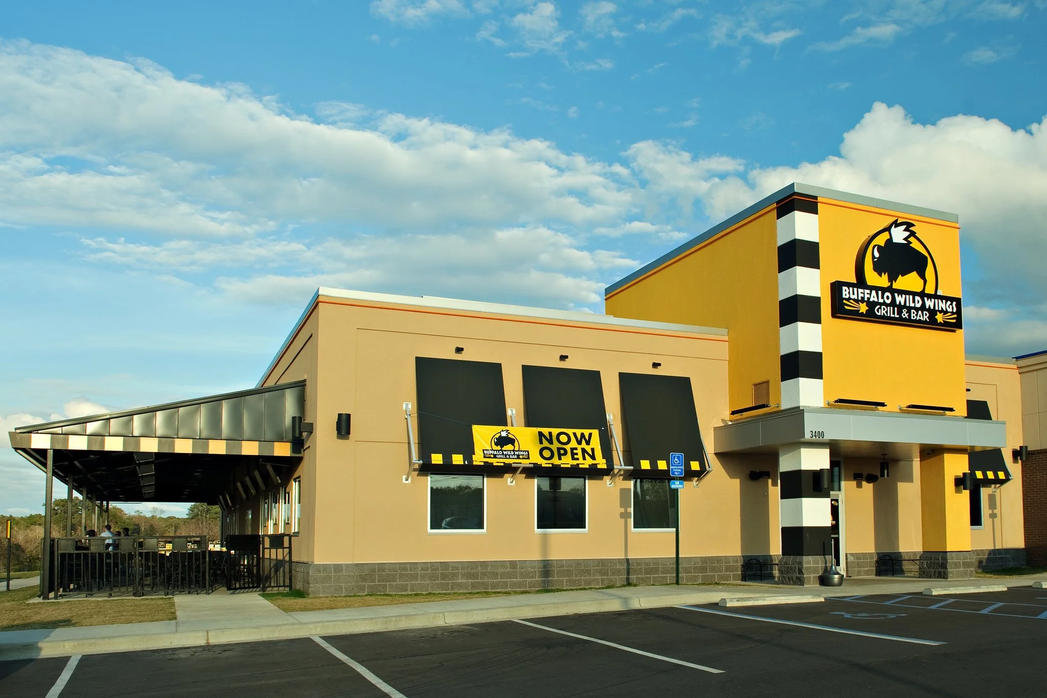 Exterior view of Buffalo Wild Wings restaurant with a yellow sign reading 'NOW OPEN' and blue sky with clouds in the background.