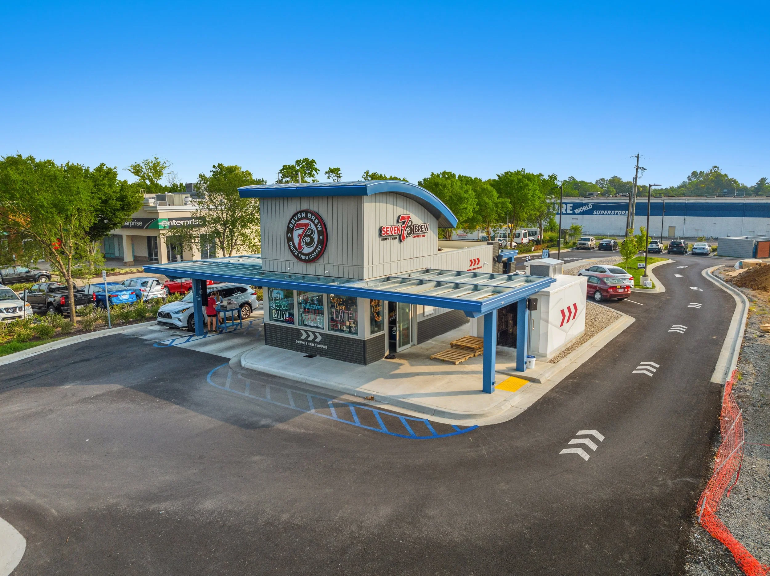 A small drive-thru coffee stand named 'Seven Brew' in a parking lot, with a blue roof and signage, a few customers, and cars parked around, with a large shopping store in the background under a clear blue sky.