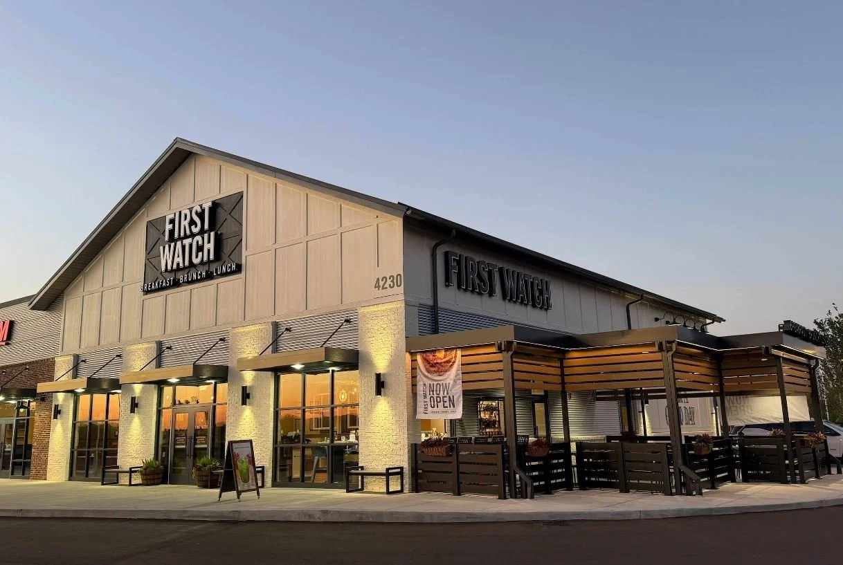 Exterior view of a First Watch restaurant at dusk with a sign indicating it is now open, featuring outdoor seating areas and illuminated entrance lights.