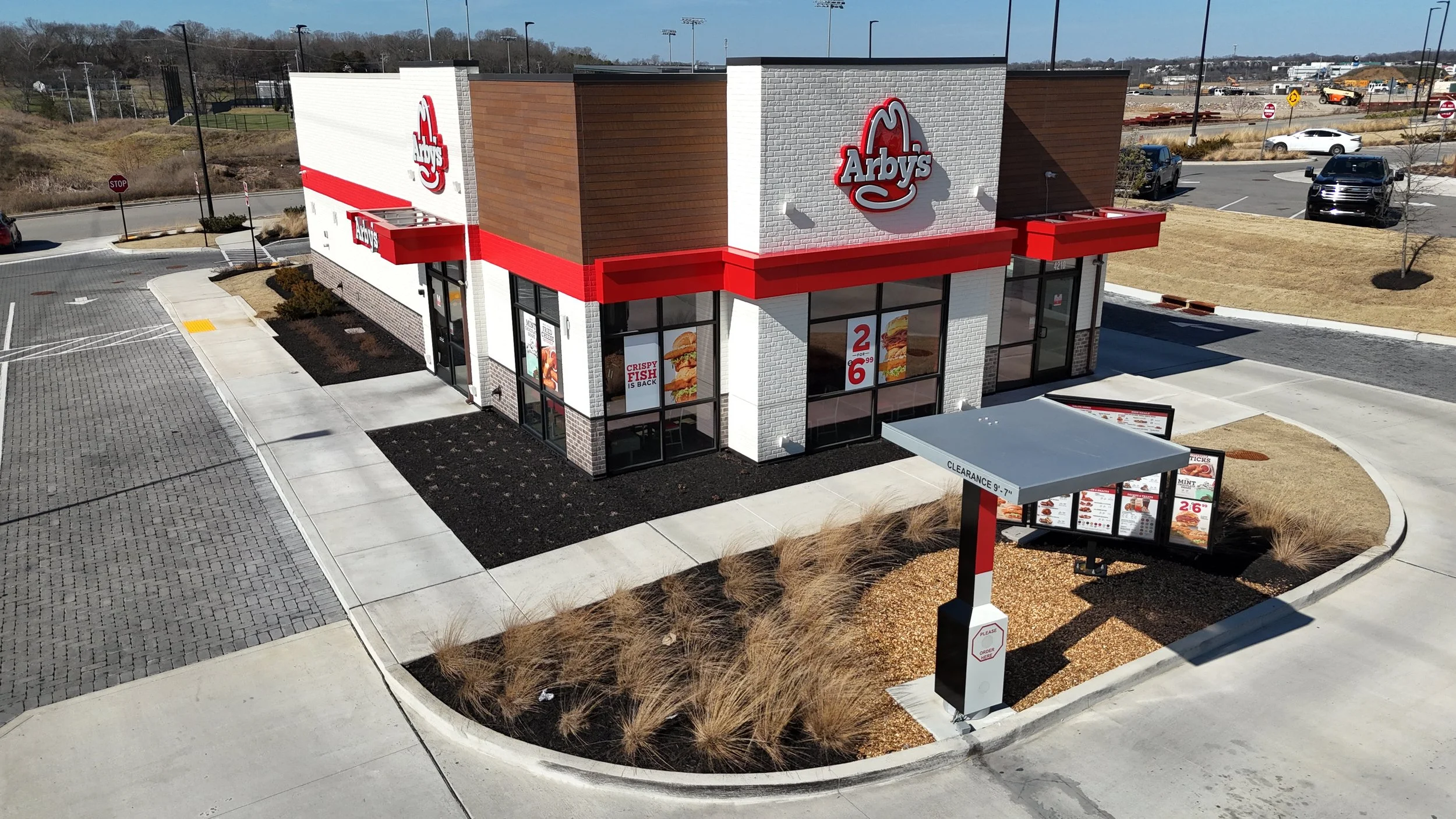 A new Arby's restaurant with a modern design, featuring white brick and wooden panel exterior, red accents, large windows, and outdoor signage, located in a parking lot on a sunny day.