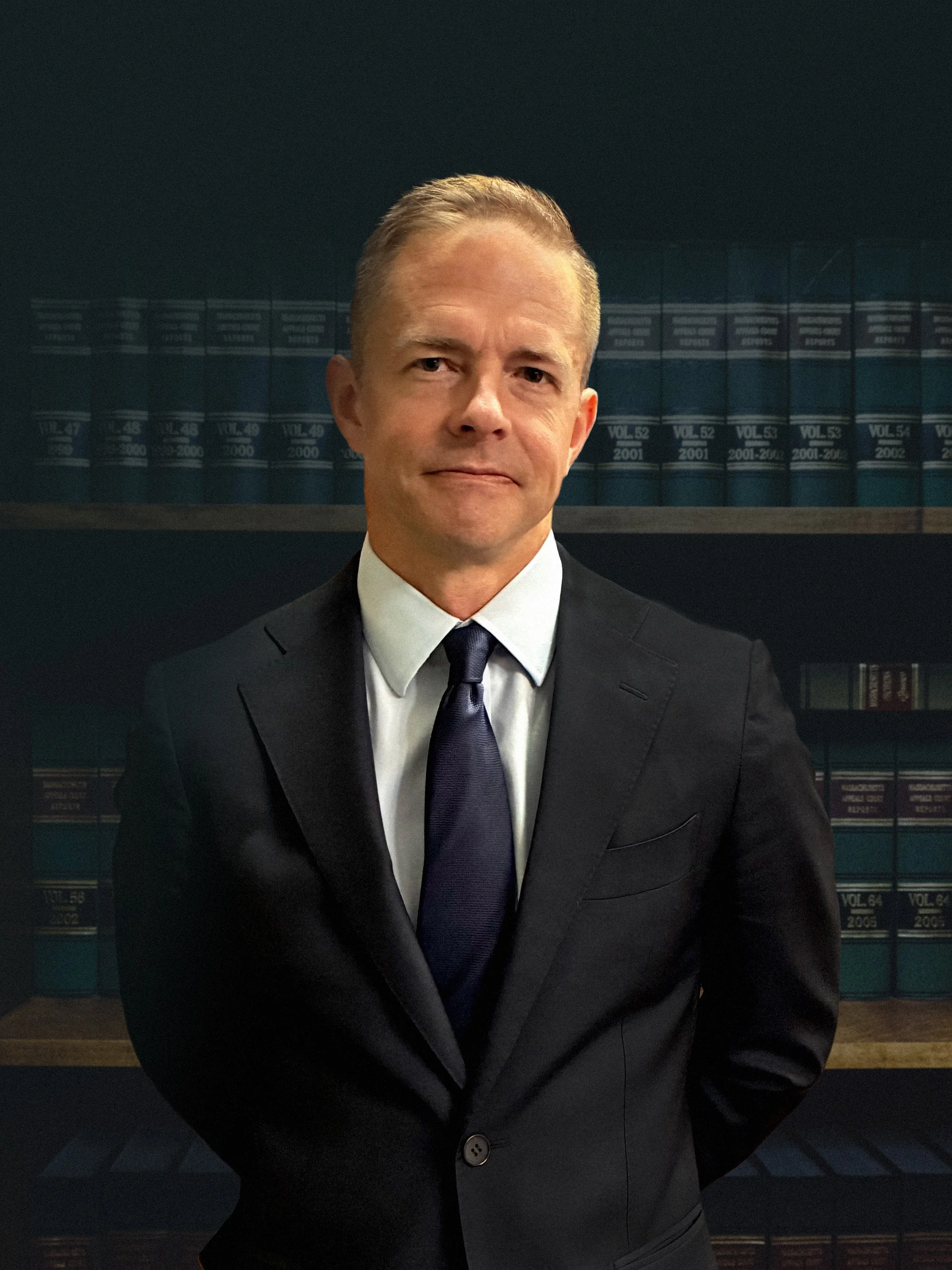 Attorney Lucas Newbill in a dark suit, white shirt, and dark tie standing in front of a bookshelf filled with law books.