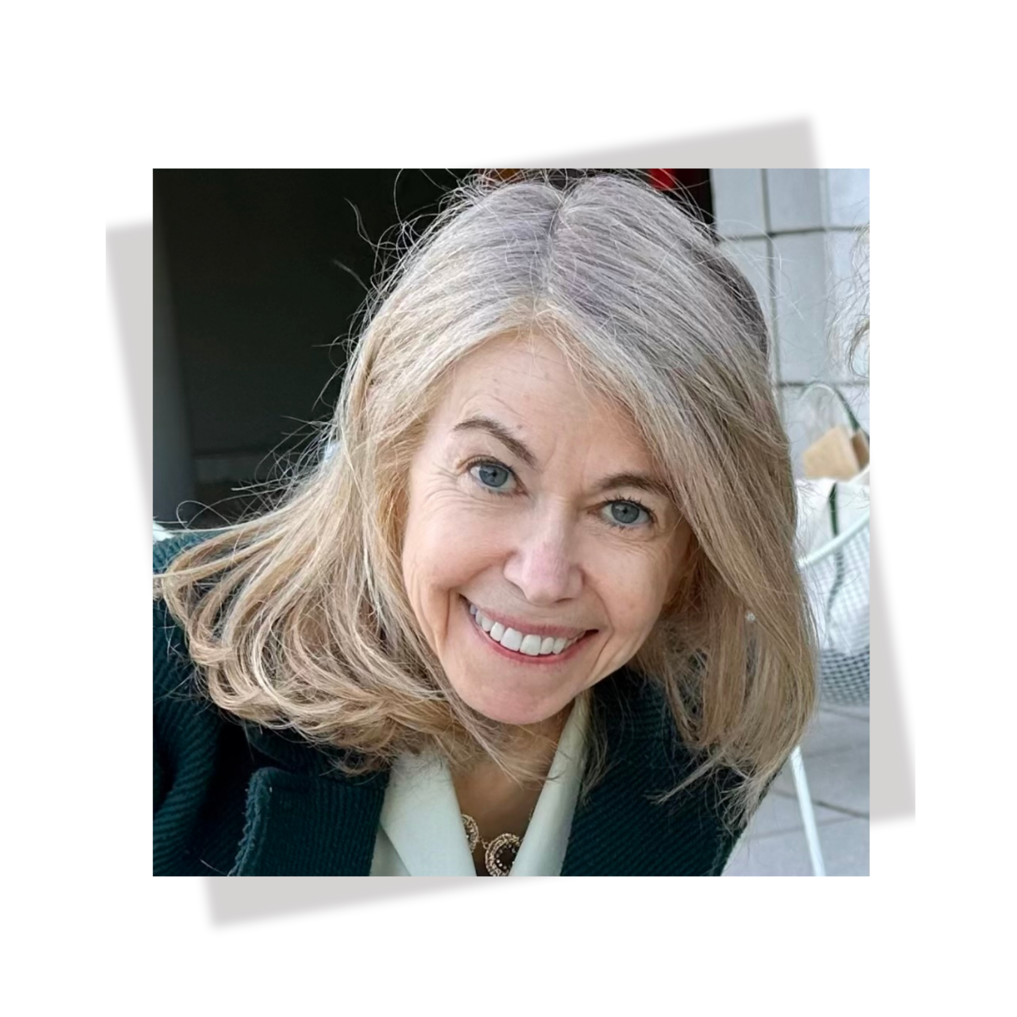Close-up photo of a woman with shoulder-length blonde hair, blue eyes, and a bright smile, wearing a dark blazer and a necklace, sitting outdoors near a tiled wall.