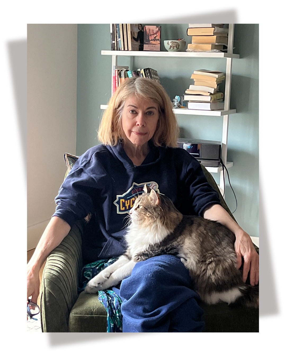 An older woman sitting on a green armchair with a large, long-haired tabby cat on her lap, in a room with bookshelves in the background.