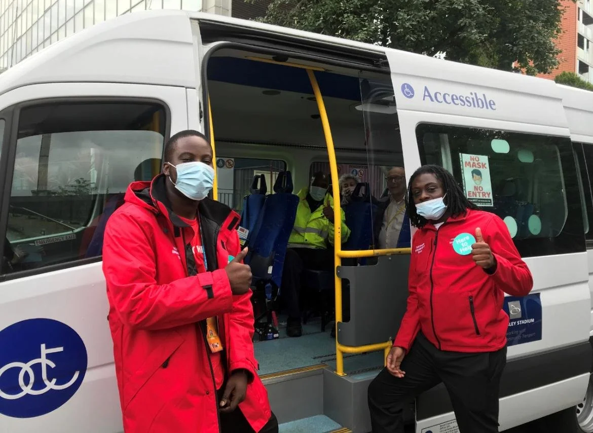 Two bus attendants wearing red jackets and face masks giving thumbs up in front of an accessible bus with passengers inside, some wearing yellow vests, during daytime.