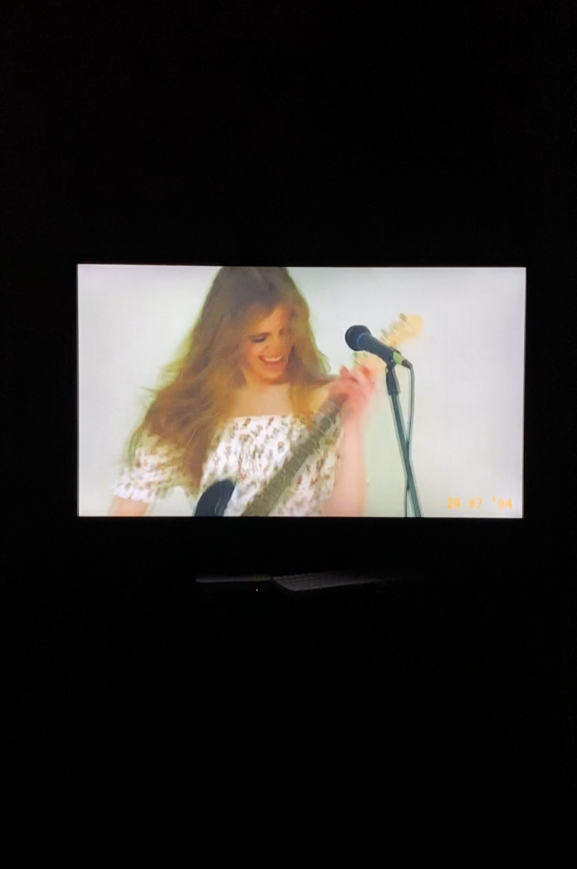 A young woman with long, wavy hair smiling while holding a guitar and standing at a microphone.