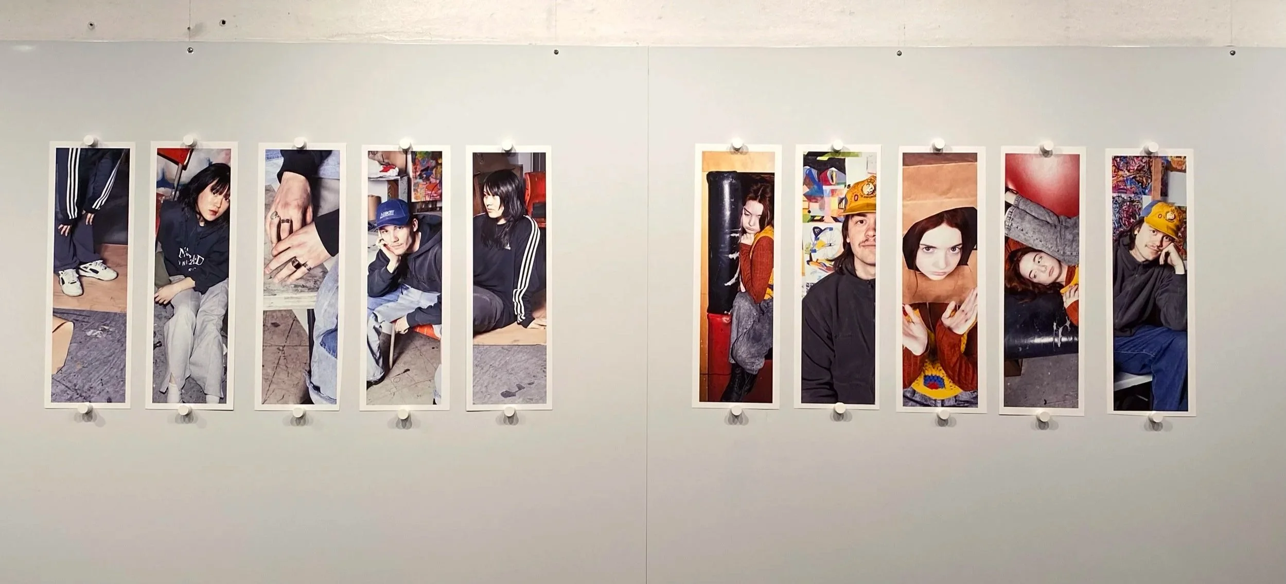 Exhibition wall displaying ten long photographic portraits of young individuals, each mounted with thumbs-up magnets.