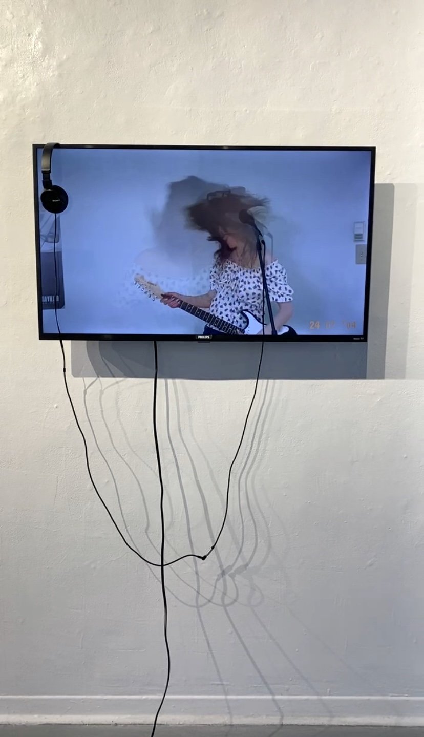 Television monitor displaying woman with curly hair playing an electric guitar, wearing a polka dot shirt, with headphones hanging on the side of the TV, and black cables hanging down and casting shadows on the wall.