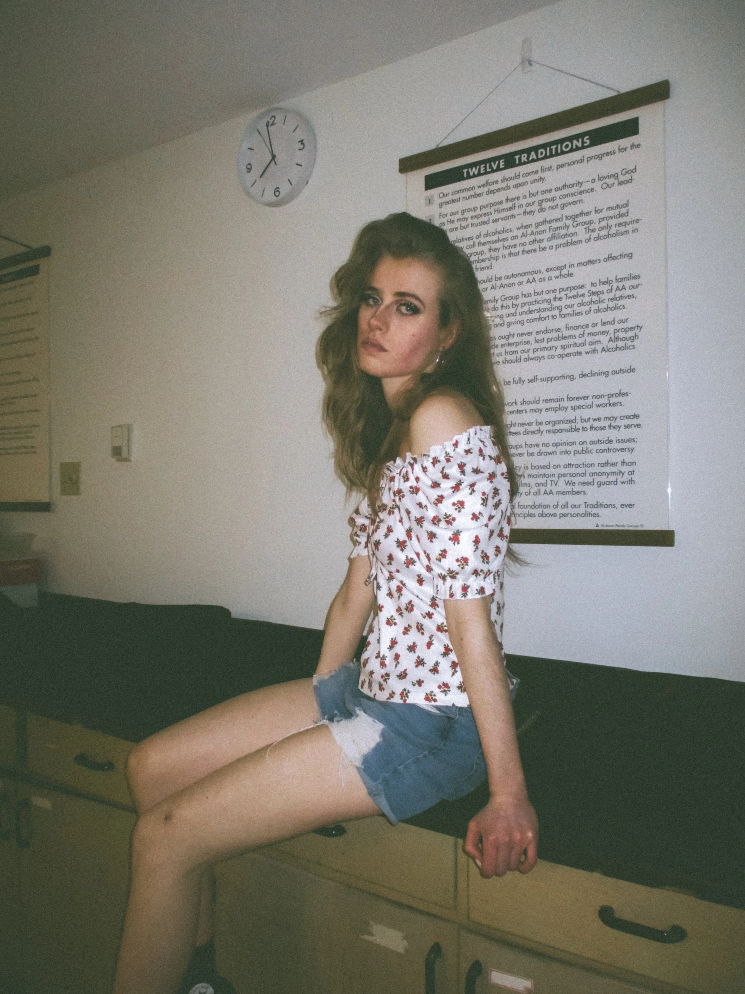 A young woman with long wavy brown hair, wearing a white off-the-shoulder top with red floral print and denim shorts, sitting on a black countertop in a room with a white wall, a wall-mounted clock showing 12:09, and a large poster titled "Twelve Tra