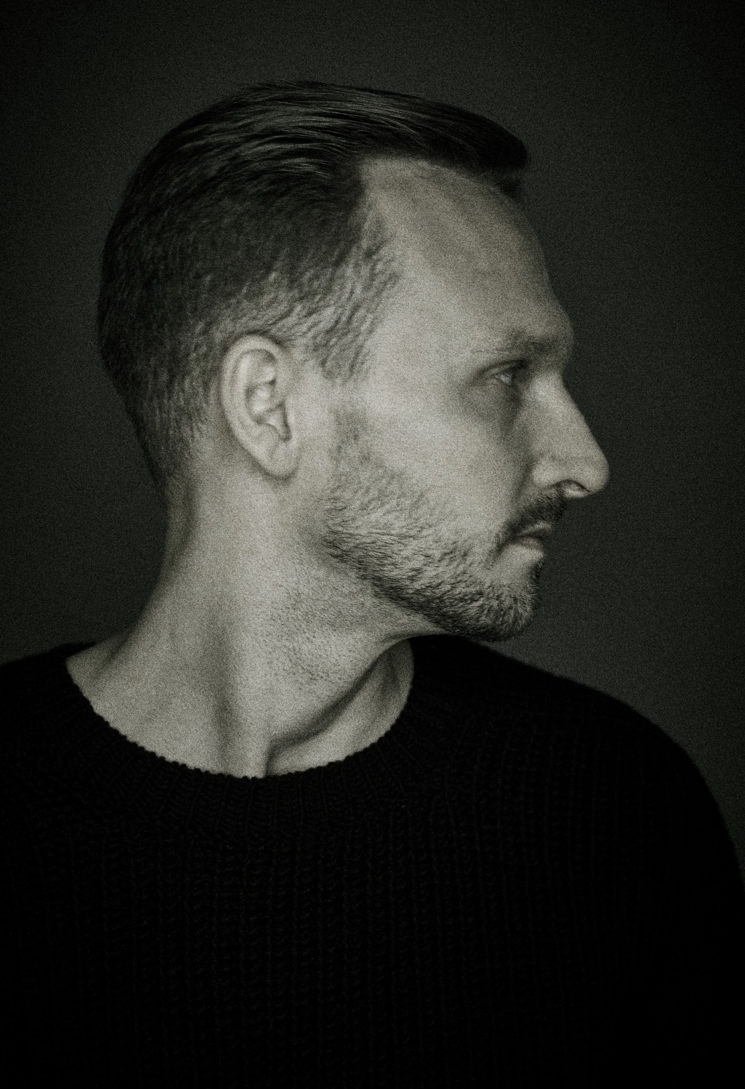 Side profile of a man with short, dark, slicked-back hair and a beard, wearing a black sweater against a dark background.