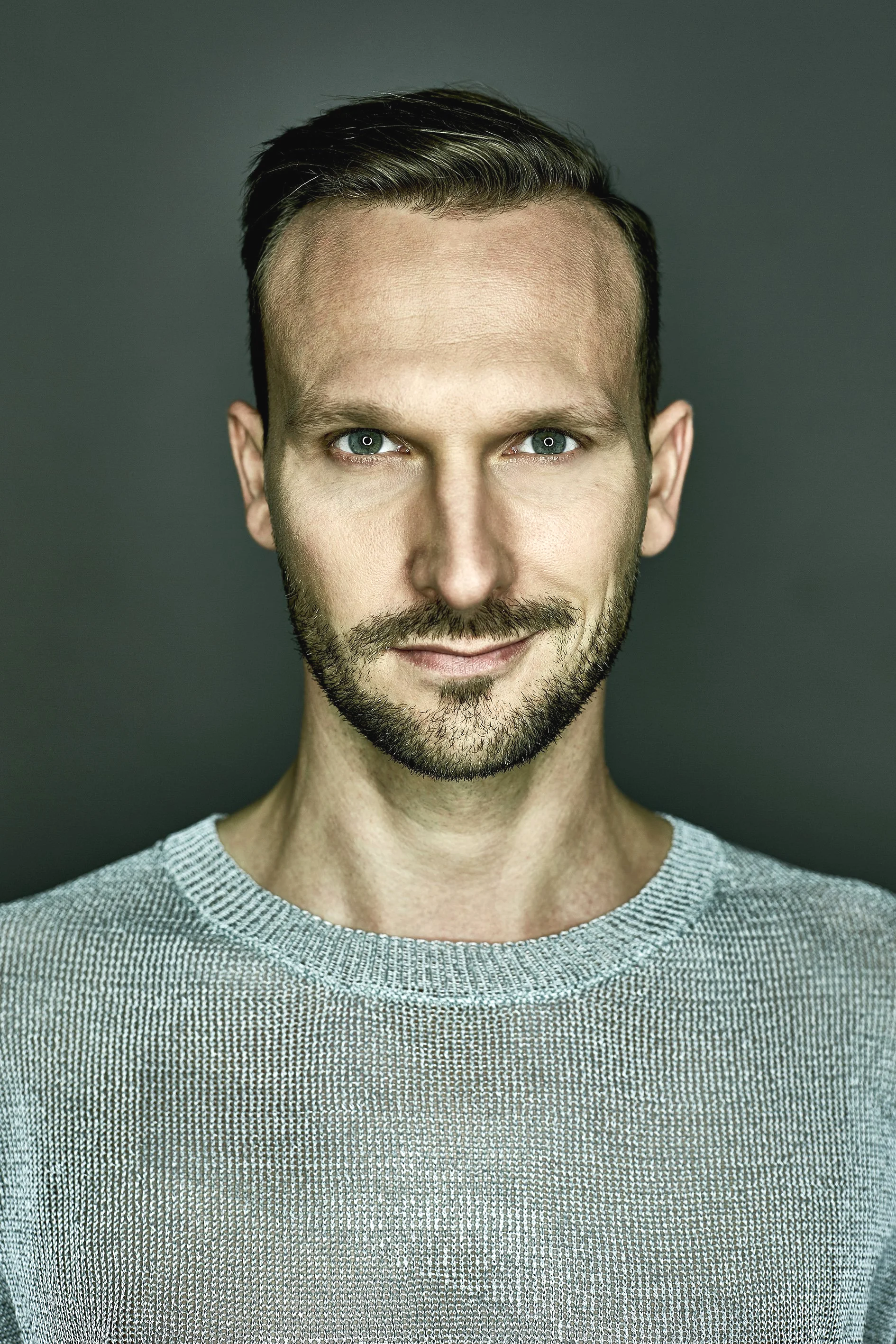 Close-up of a man with short brown hair, a beard, and blue eyes, wearing a gray textured sweater, smiling slightly, against a dark background.