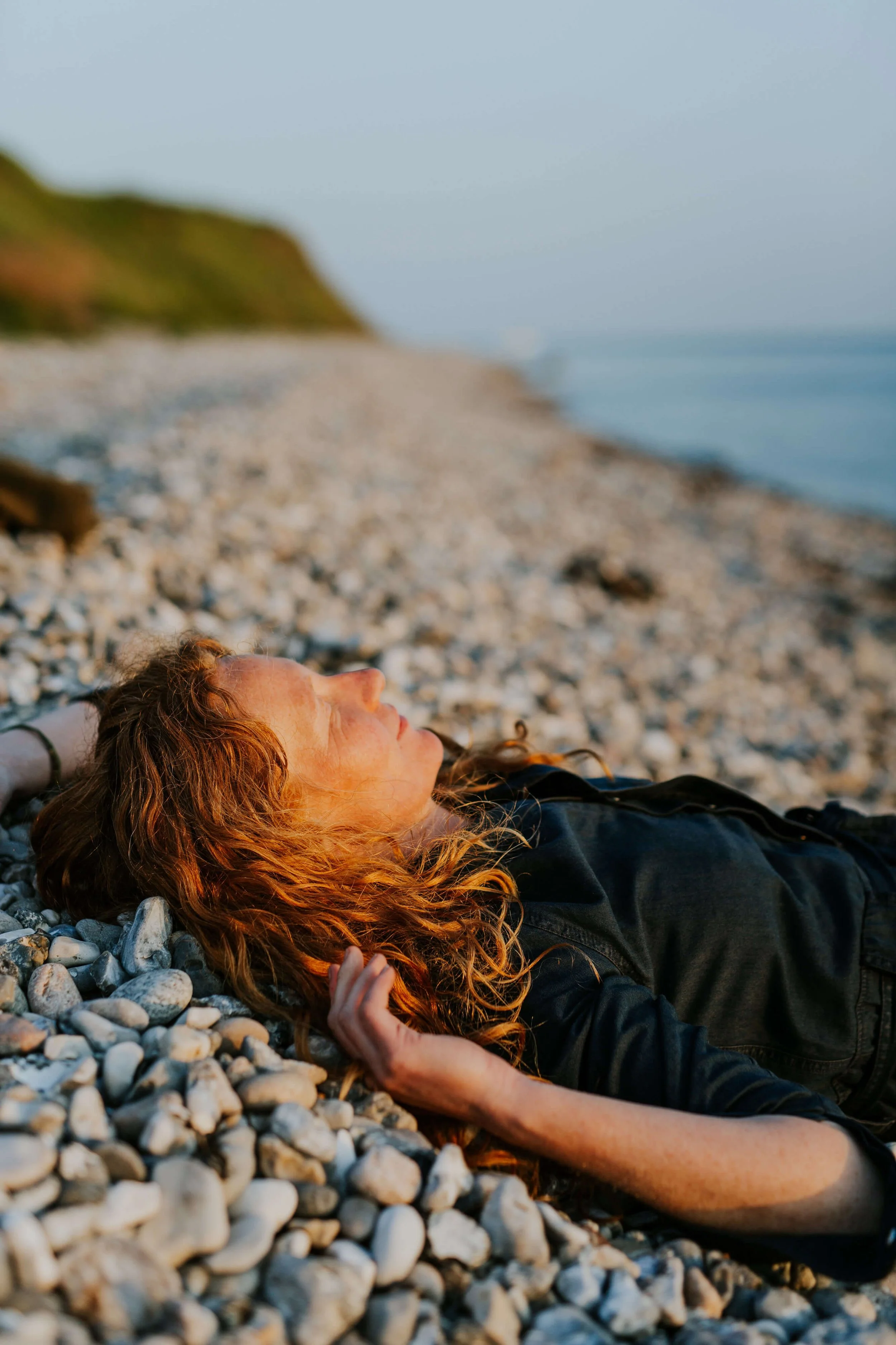 Kvinde med langt rødt krøllet hår ligger med lukkede øjne og ansigtet mod solen på en stenet strand