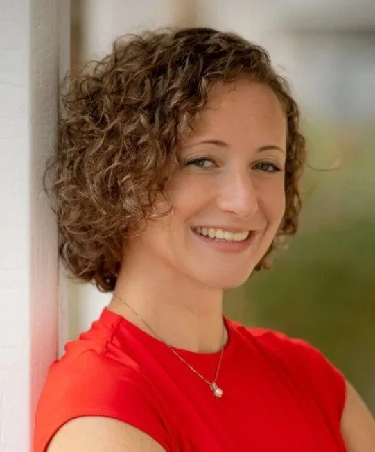 A woman with short curly brown hair smiling while leaning against a white wall, wearing a bright red top and a delicate necklace, outdoors with blurred natural background.