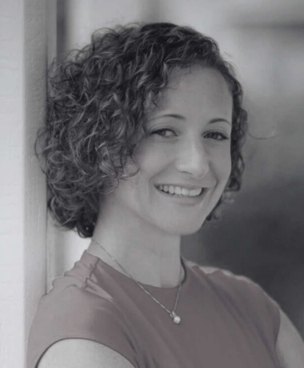 Black and white photo of a woman with short curly hair smiling and leaning against a wall.