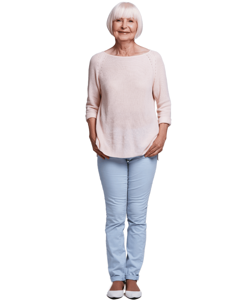 Een oudere vrouw met wit haar, gekleed in een lichtroze trui, lichtblauwe broek en witte schoenen, staand en glimlachend.