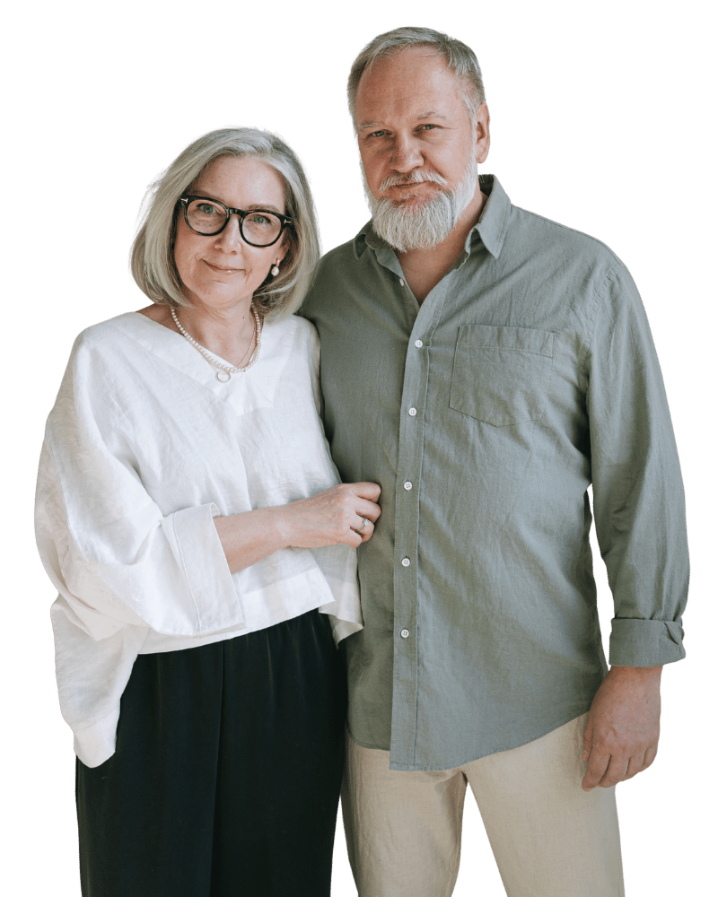 Een oudere vrouw en man staan samen, de vrouw draagt een witte blouse en een ketting, de man draagt een grijze overhemd, beiden kijken naar de camera.