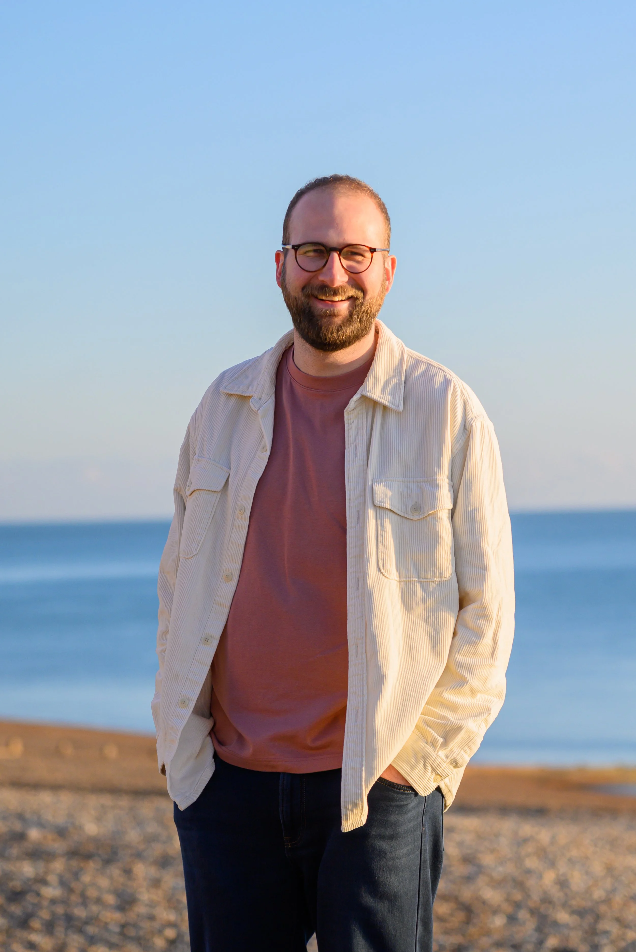 A smiling man with glasses and a beard standing outdoors on a beach with the ocean in the background during sunset.