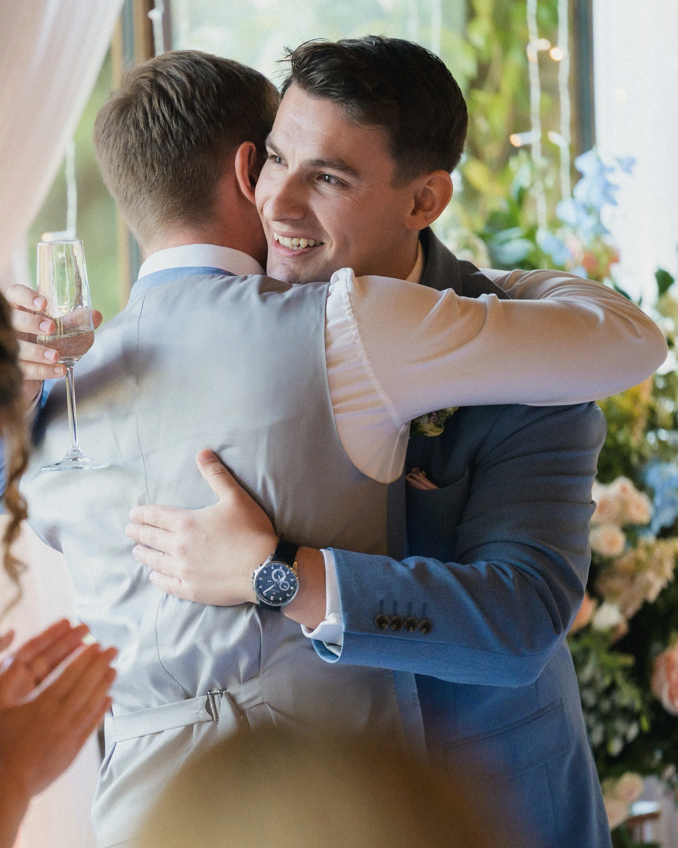 Two men embracing each other at a wedding, one holding a champagne glass, with a blurred background of plants and string lights.