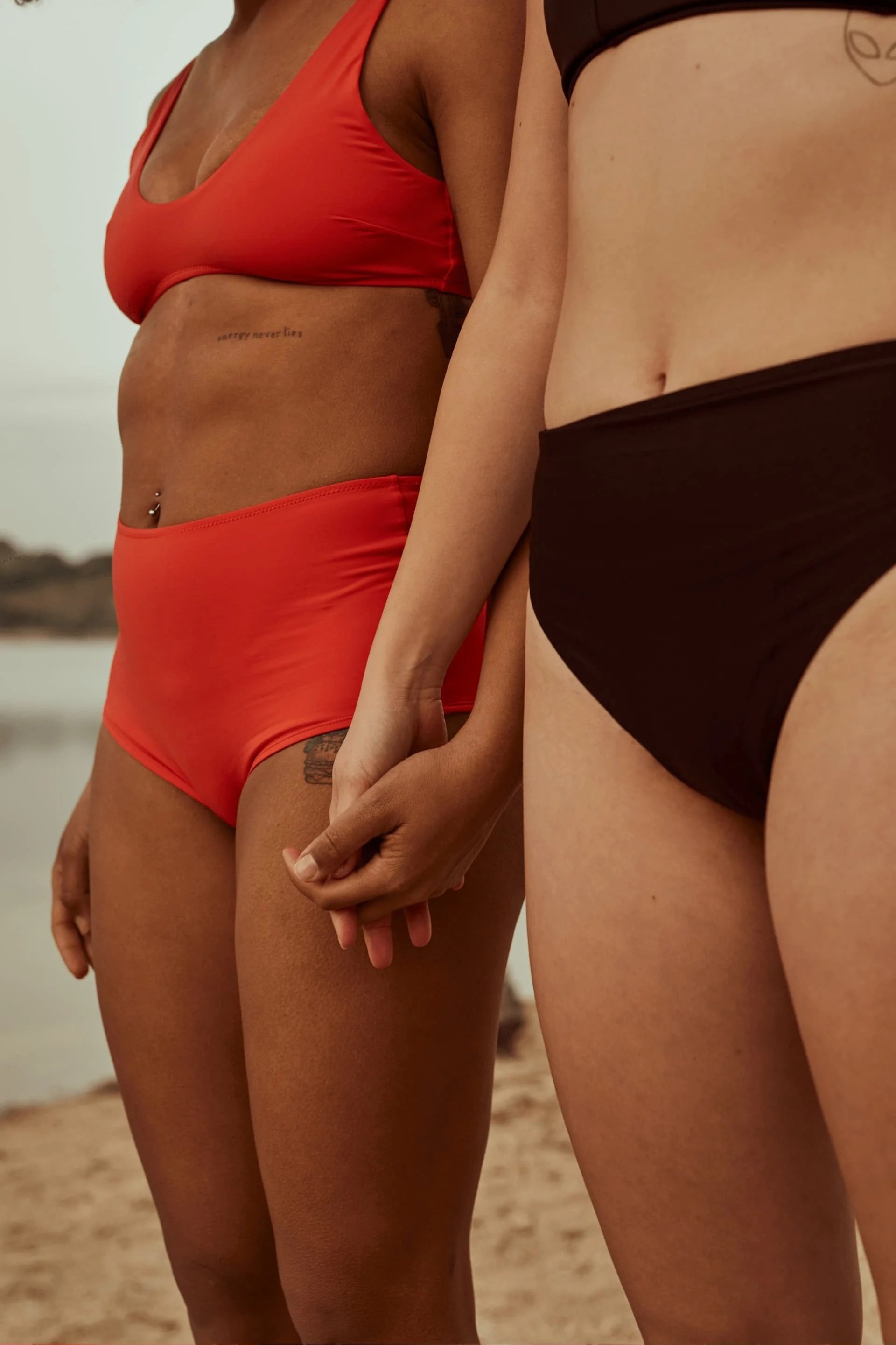 Two women standing on a beach, holding hands, wearing swimsuits, with a body of water in the background.