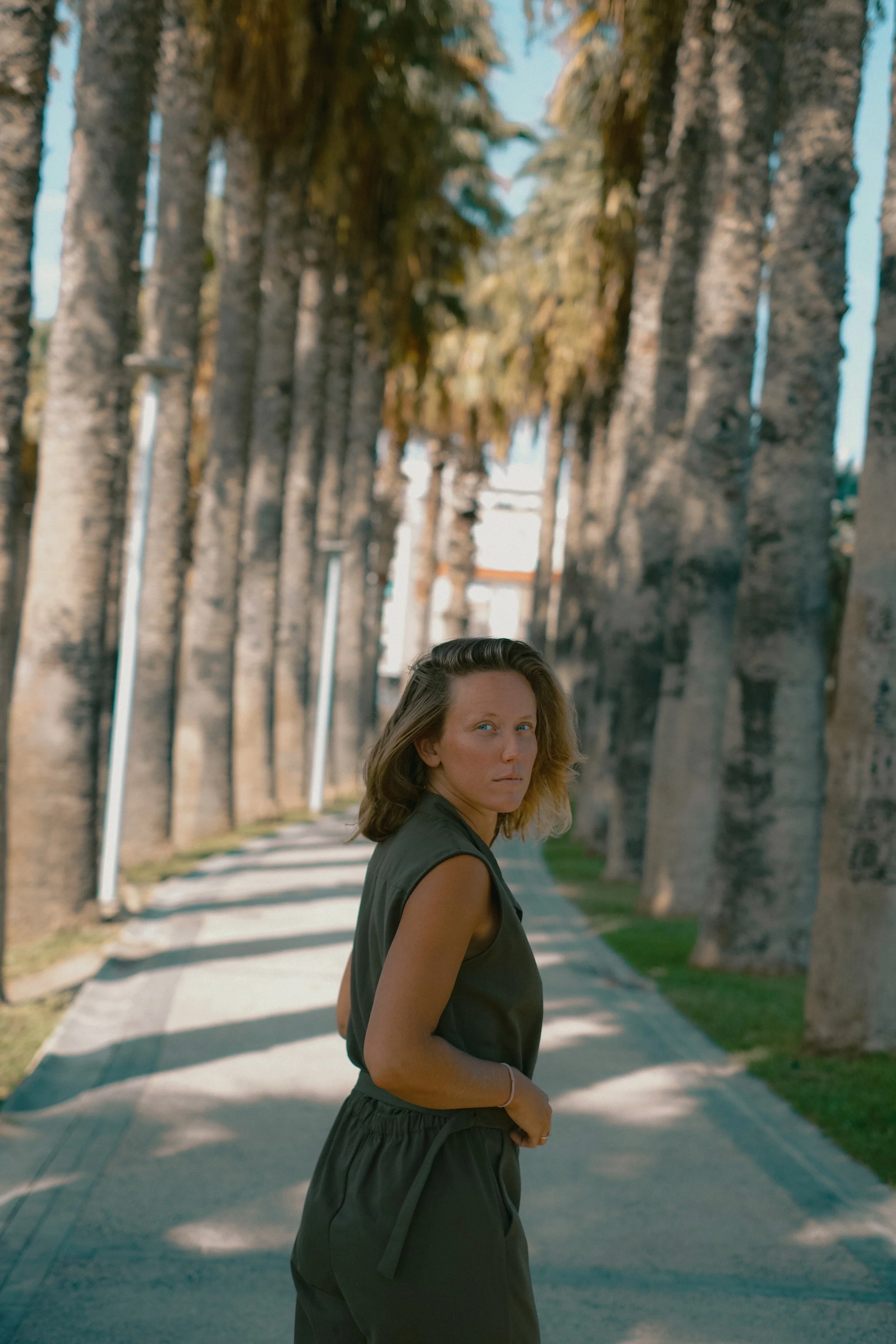 A woman with shoulder-length wavy hair standing on a tree-lined sidewalk, looking over her shoulder at the camera.