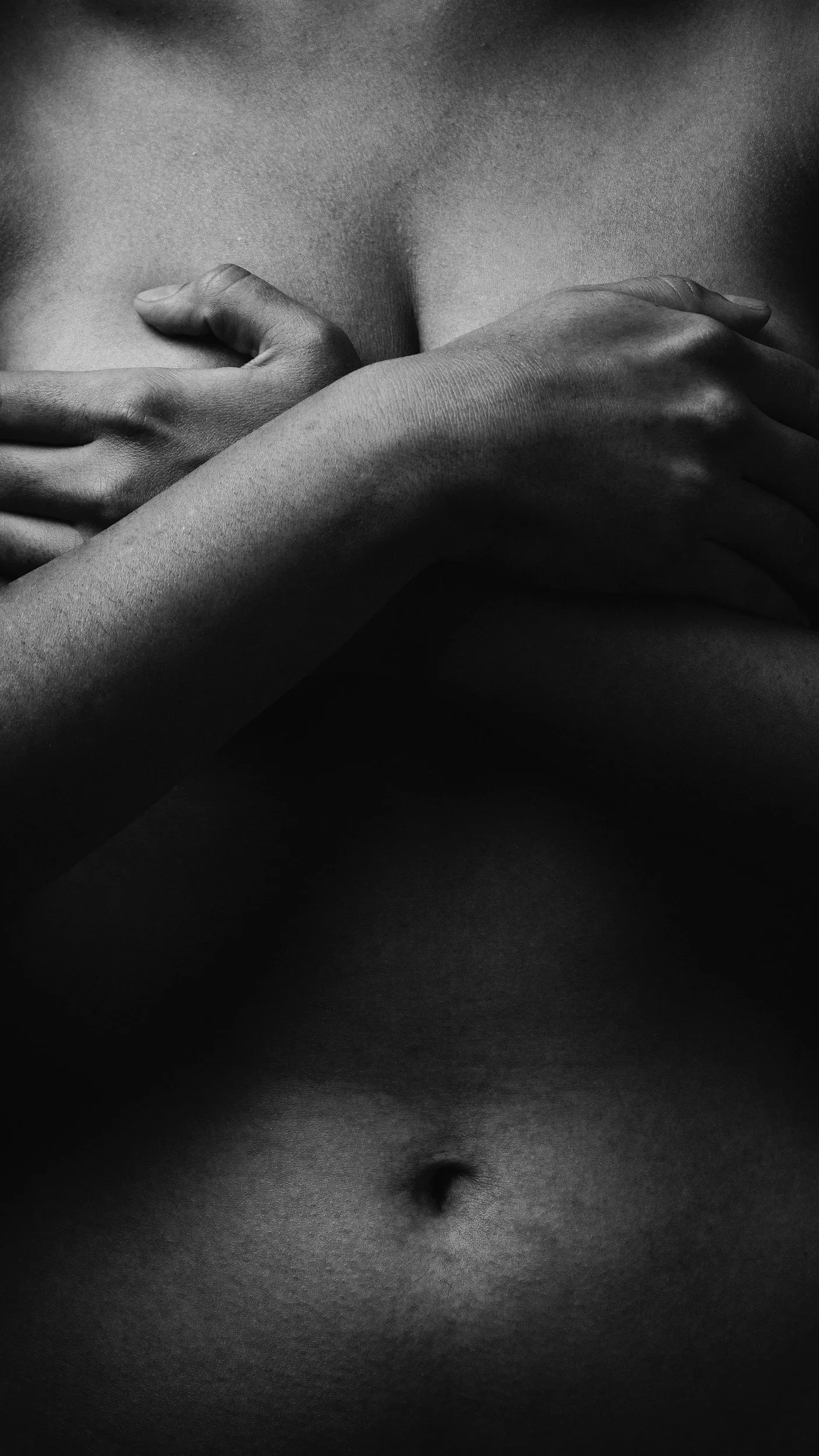 Close-up black and white photograph of a person's bare torso, featuring hands crossing over the chest and the navel visible at the bottom of the image.