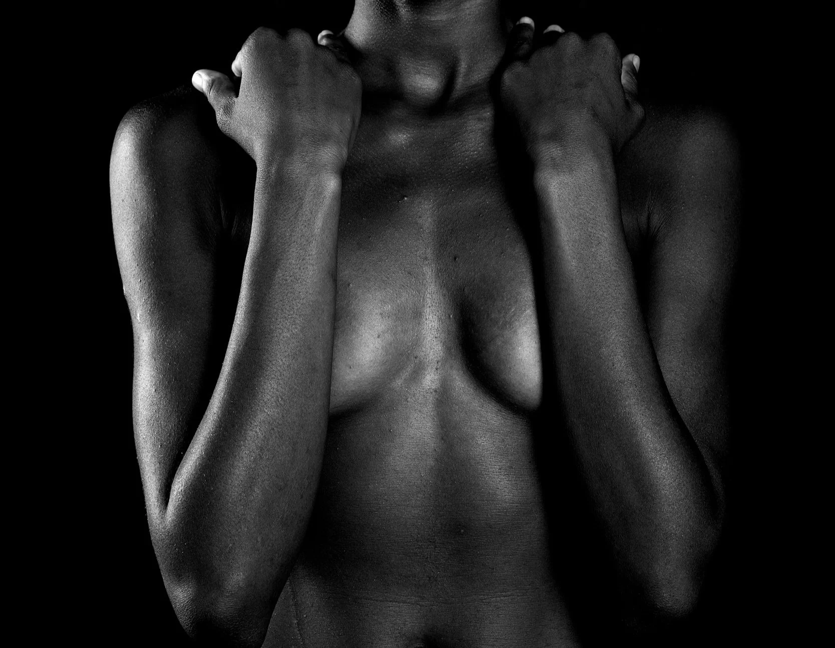 Black and white photograph of a topless woman holding her fists at her neck, with visible shoulder and arm muscles, against a dark background.