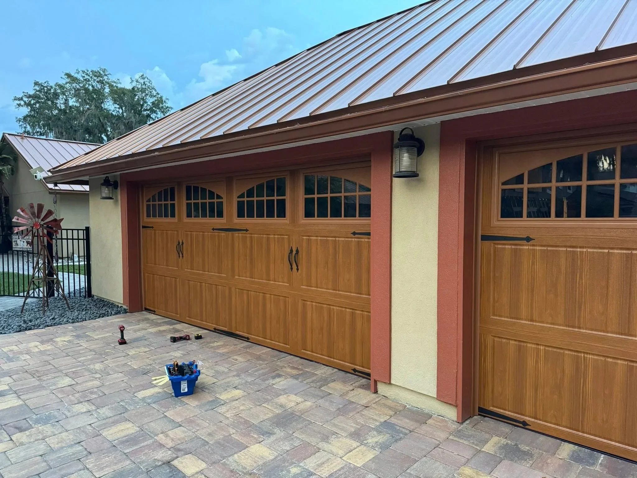 New wooden garage doors being installed on a house with a brick driveway, tools and a blue toolbox on the ground, and decorative metal wall lanterns.