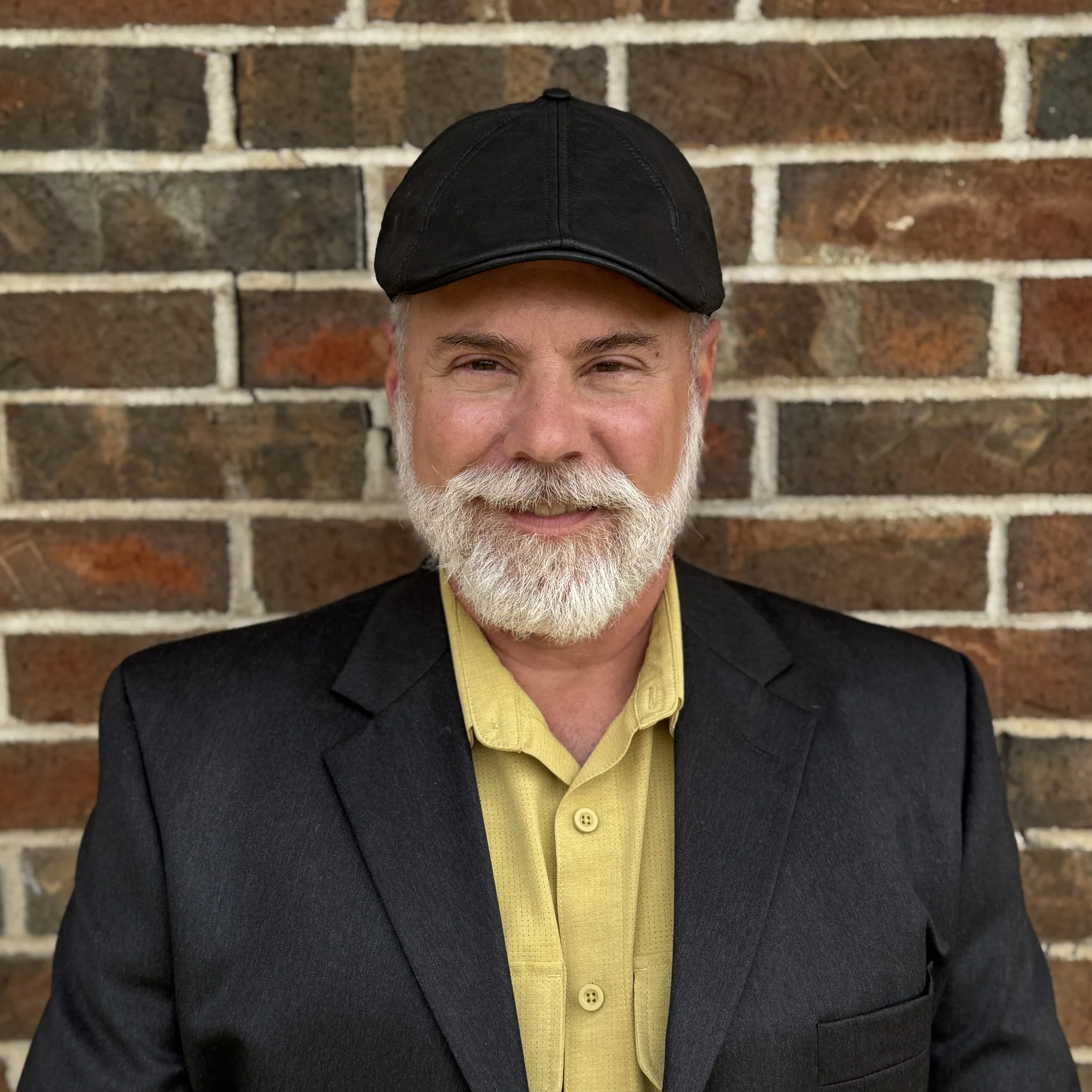 A middle-aged man with a white beard and gray hair, wearing a black cap, black blazer, and yellow shirt, standing in front of a brick wall.