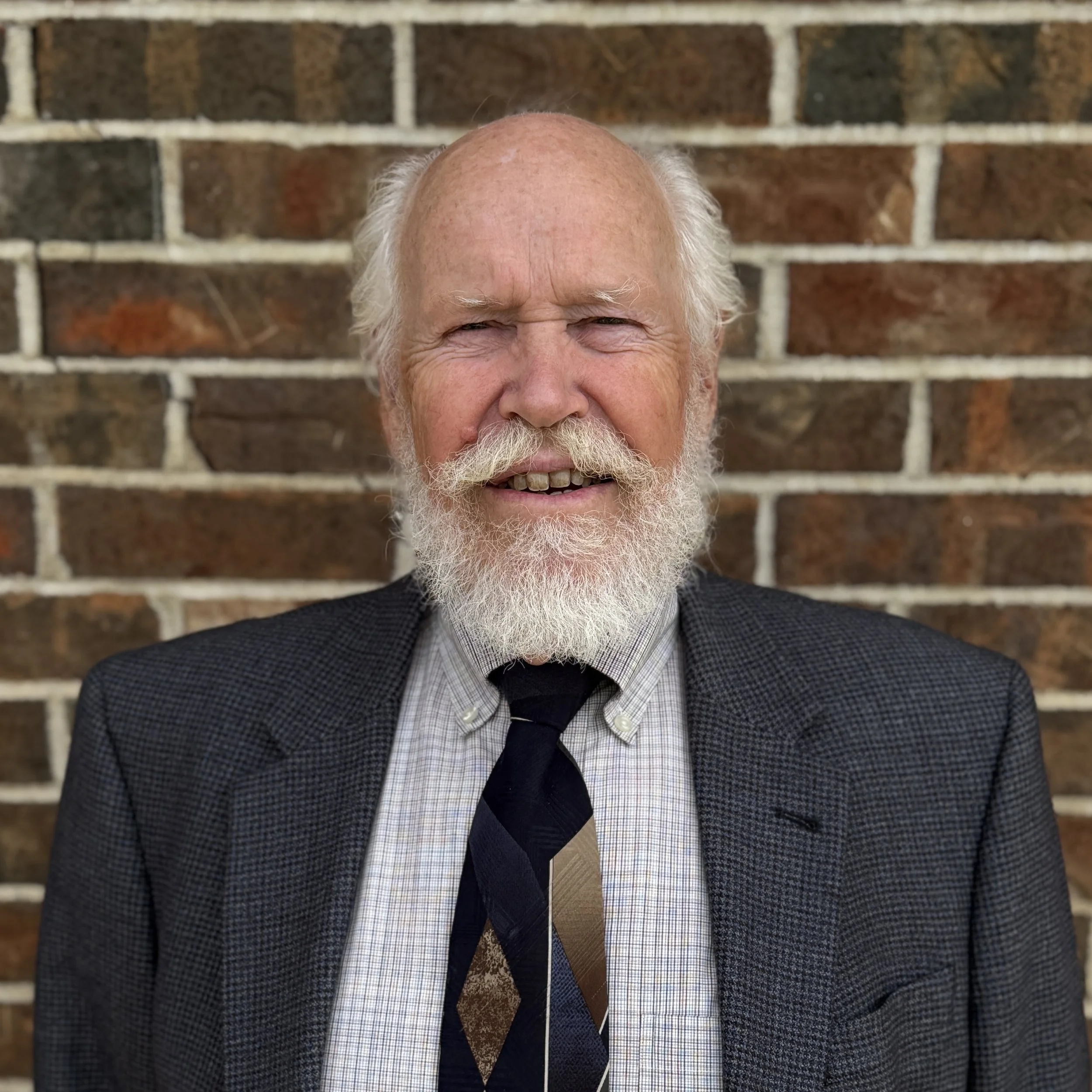 An elderly man with a white beard and mustache, wearing a checked shirt, a patterned tie, and a dark blazer, standing in front of a brick wall.