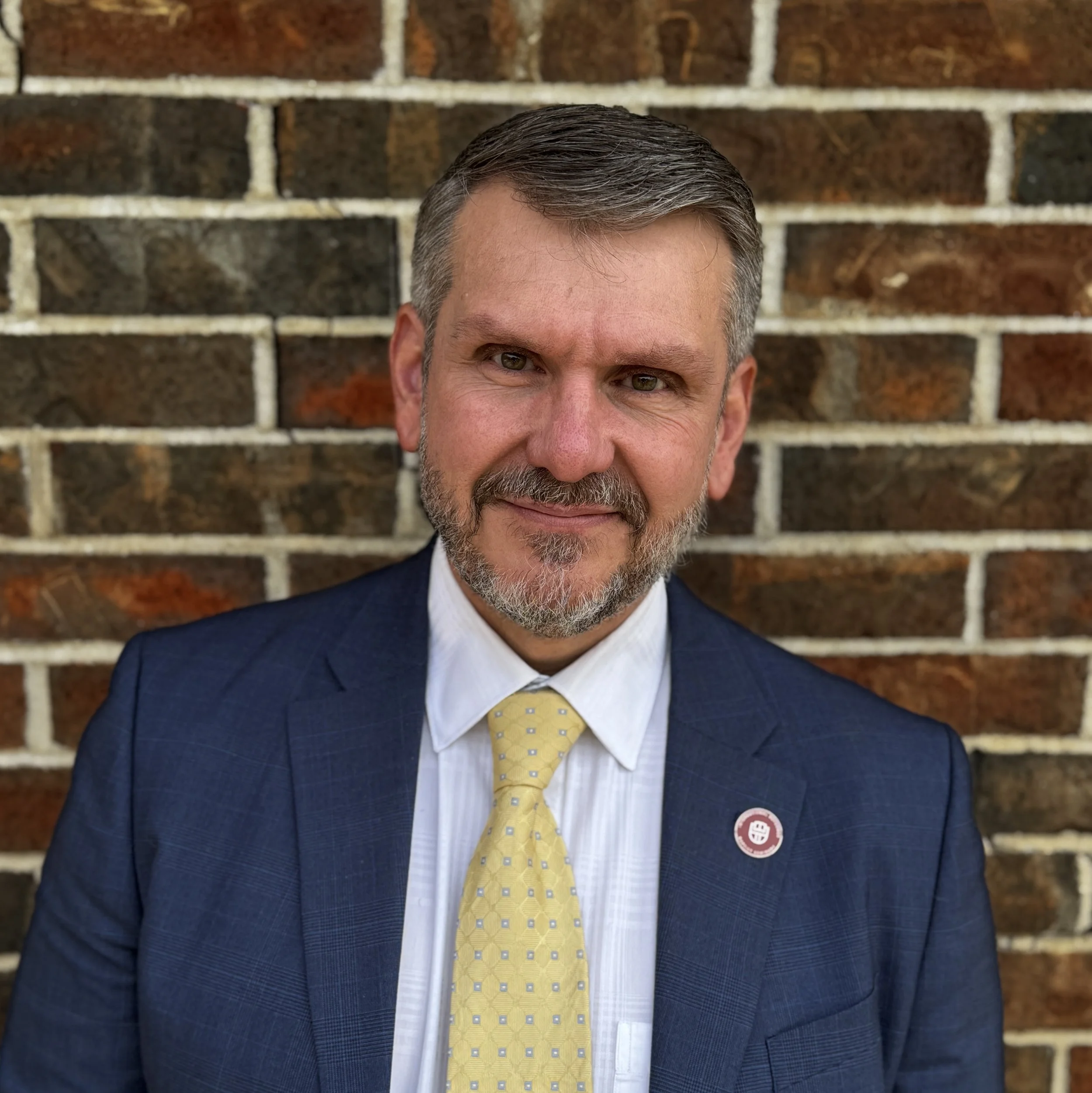 A middle-aged man with a beard and short gray hair wearing a navy blue suit, white dress shirt, and yellow polka-dot tie, standing in front of a brick wall.