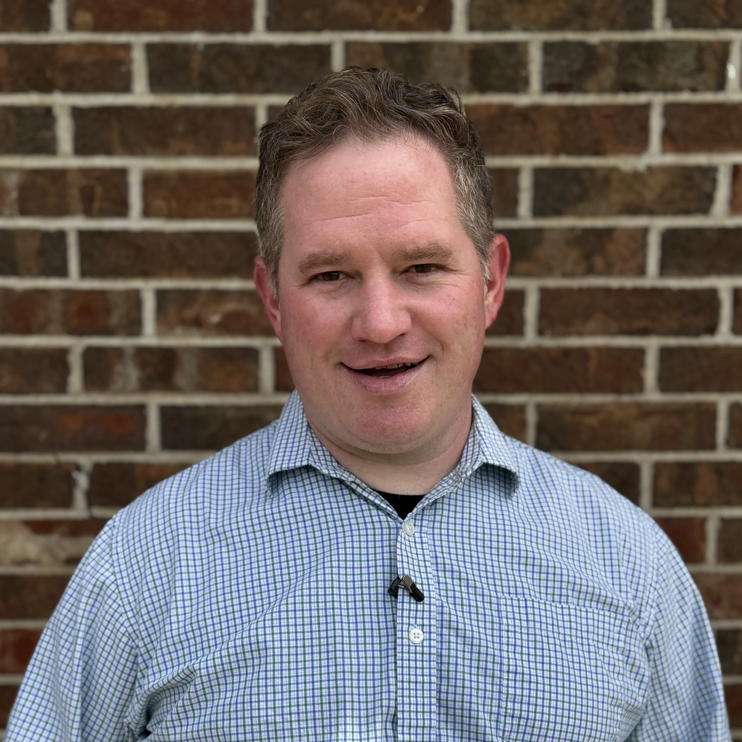 A man with short light brown hair, wearing a light blue checkered shirt, standing outdoors in front of a brick wall.