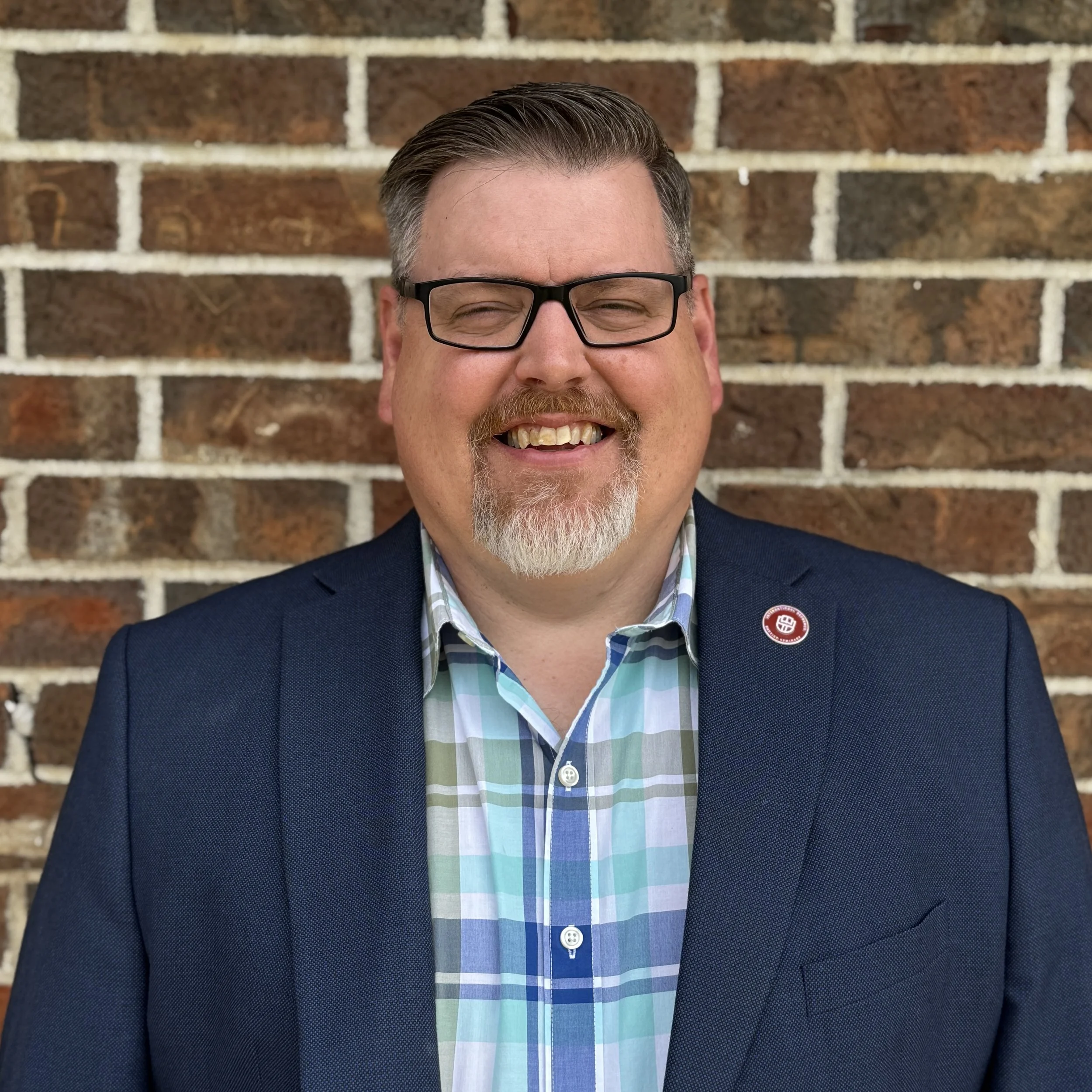 A smiling man in glasses wearing a blue blazer and a plaid shirt, standing in front of a brick wall.