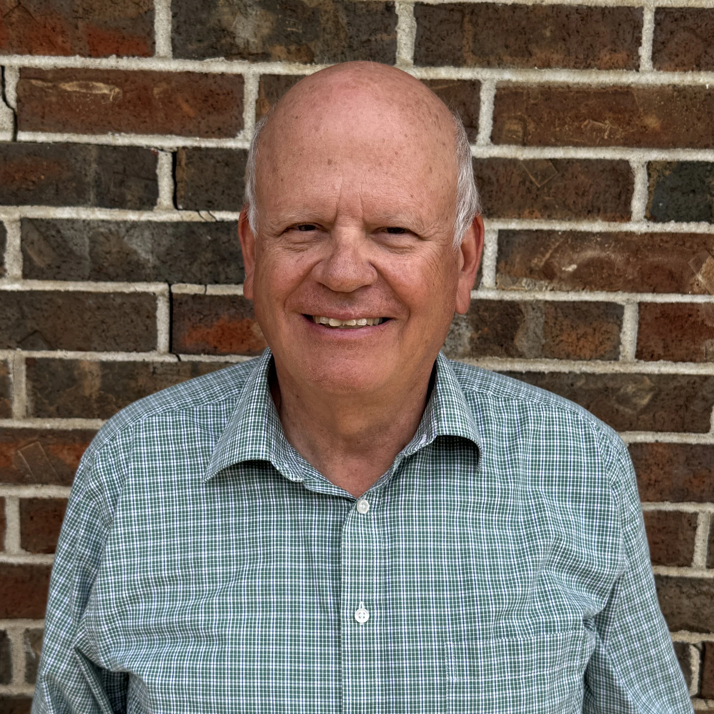 A smiling older man with a bald head and light skin, wearing a green and white checkered shirt, standing in front of a brick wall.