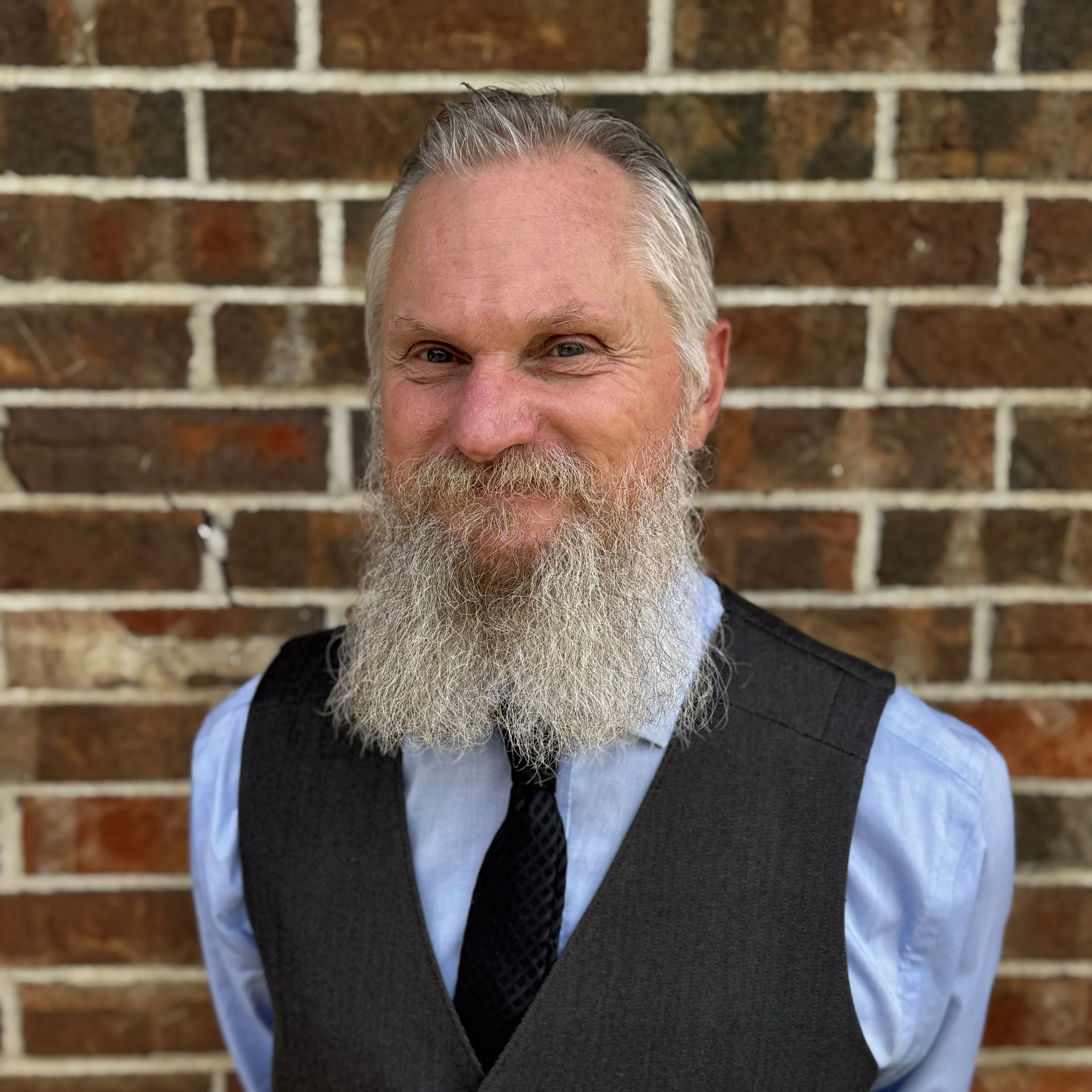 A middle-aged man with a full gray beard, gray hair, wearing a light blue shirt, a black vest, and a black tie, smiling in front of a brick wall.
