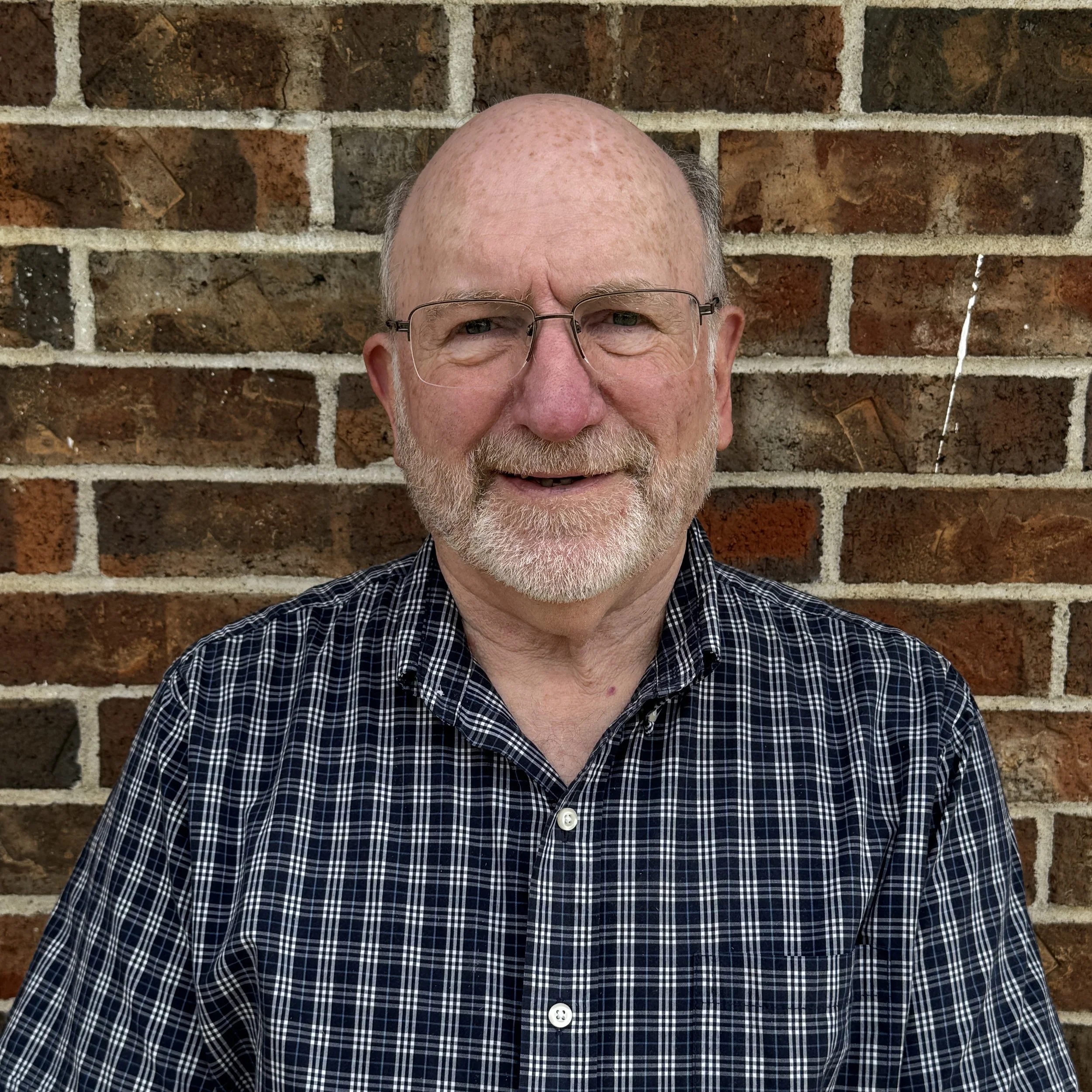 A portrait of an elderly man with glasses, a beard, and a bald head, smiling in front of a brick wall.