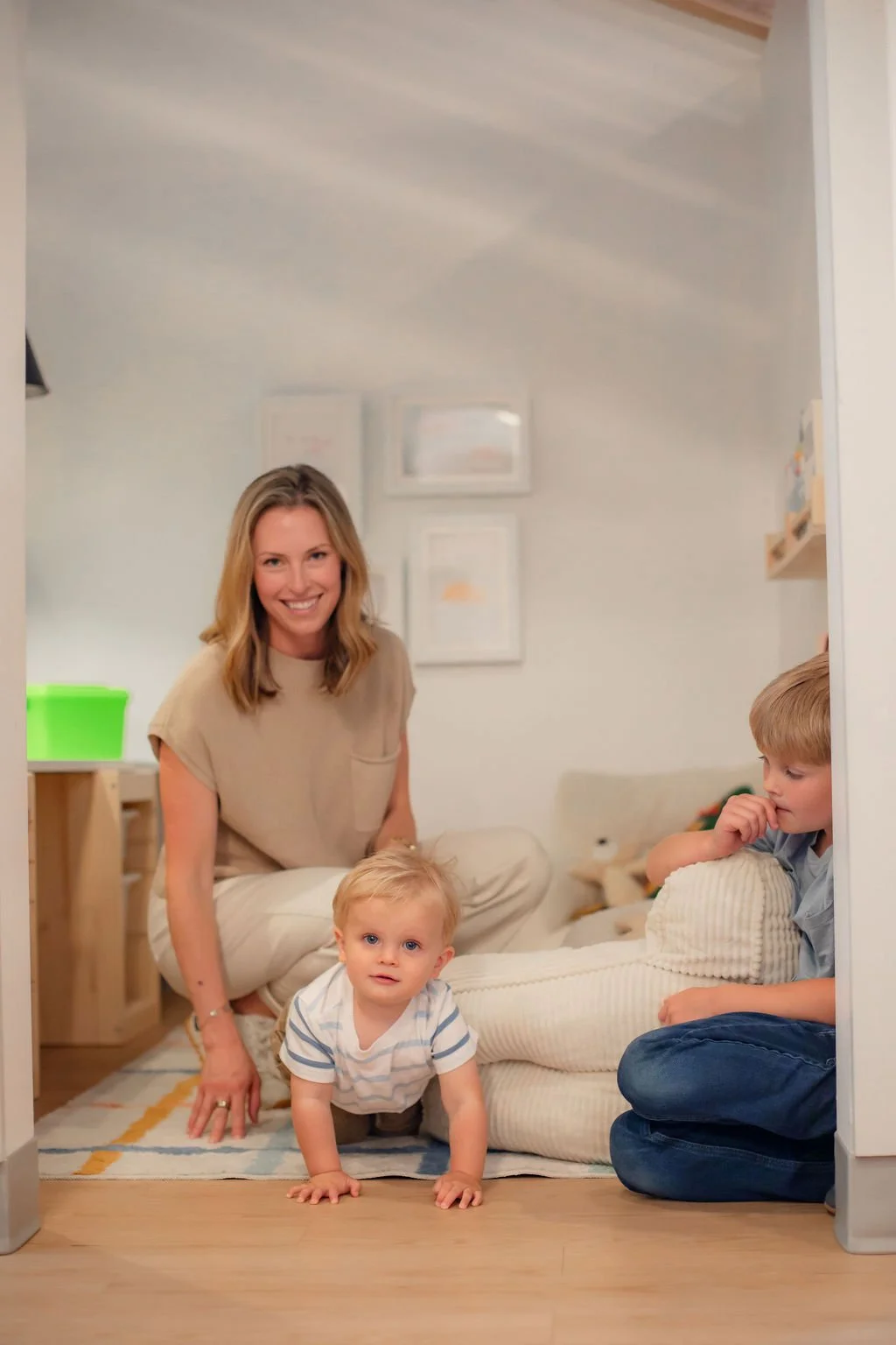 A woman with blonde hair smiling on a rug with two young boys—one crawling and one sitting and thinking—inside a cozy, well-lit room decorated with framed art.