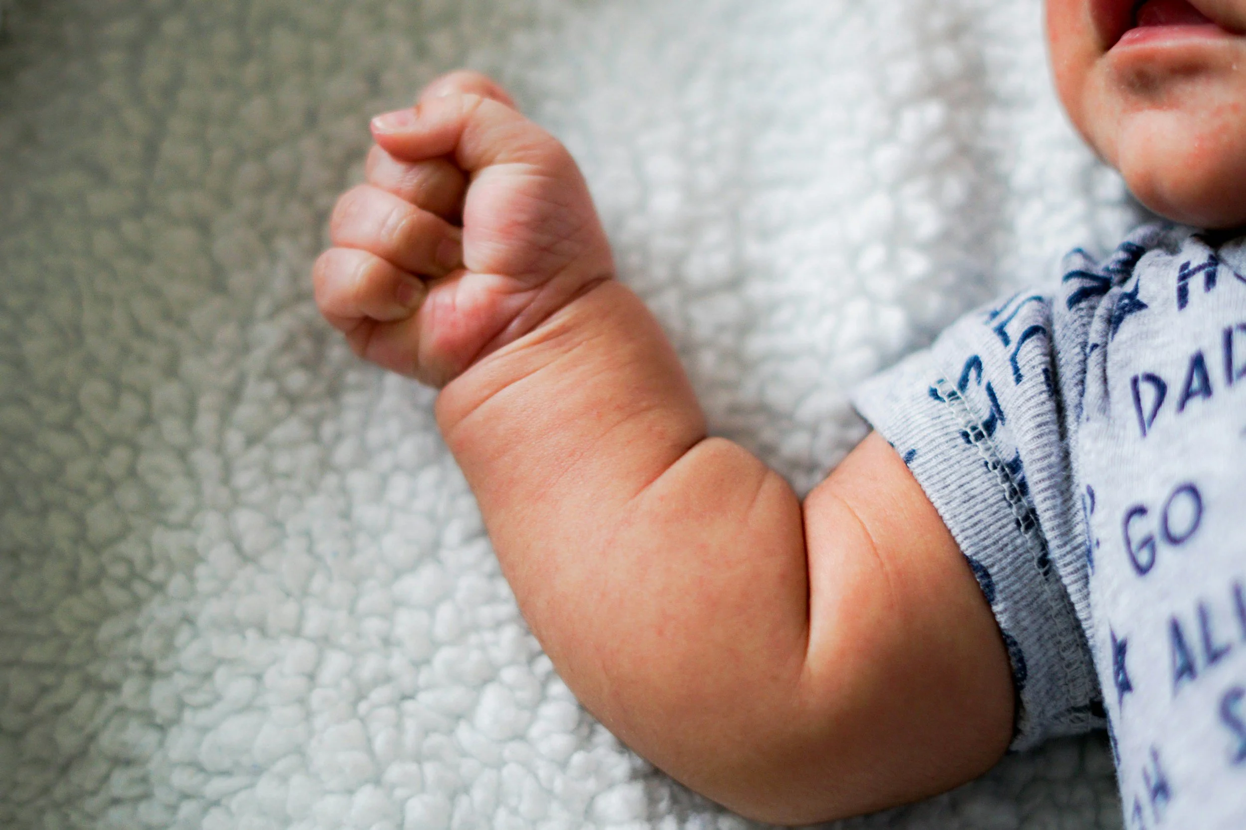 Close-up of a baby's hand clenched into a fist, lying on a textured white blanket, with part of baby's face and arm with a gray and blue patterned shirt visible.