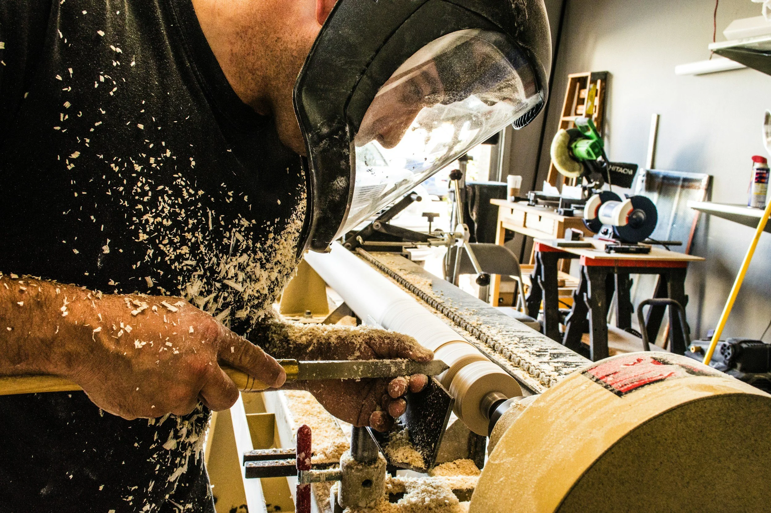 A person wearing safety goggles and a black shirt is using a wood lathe to shape a piece of wood in a workshop, with wood shavings flying around.