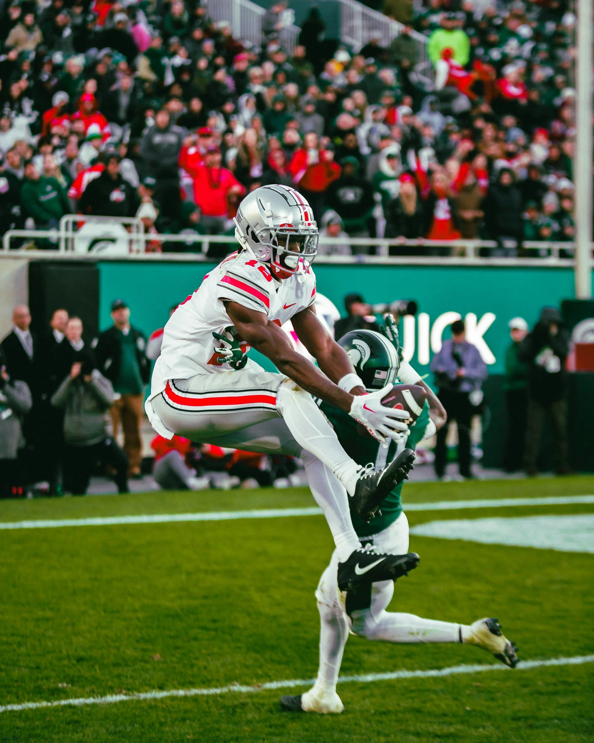 Football player in a white uniform with red and black accents catching the ball in the end zone while a defender in green attempts to block him, with a packed stadium of spectators in the background.