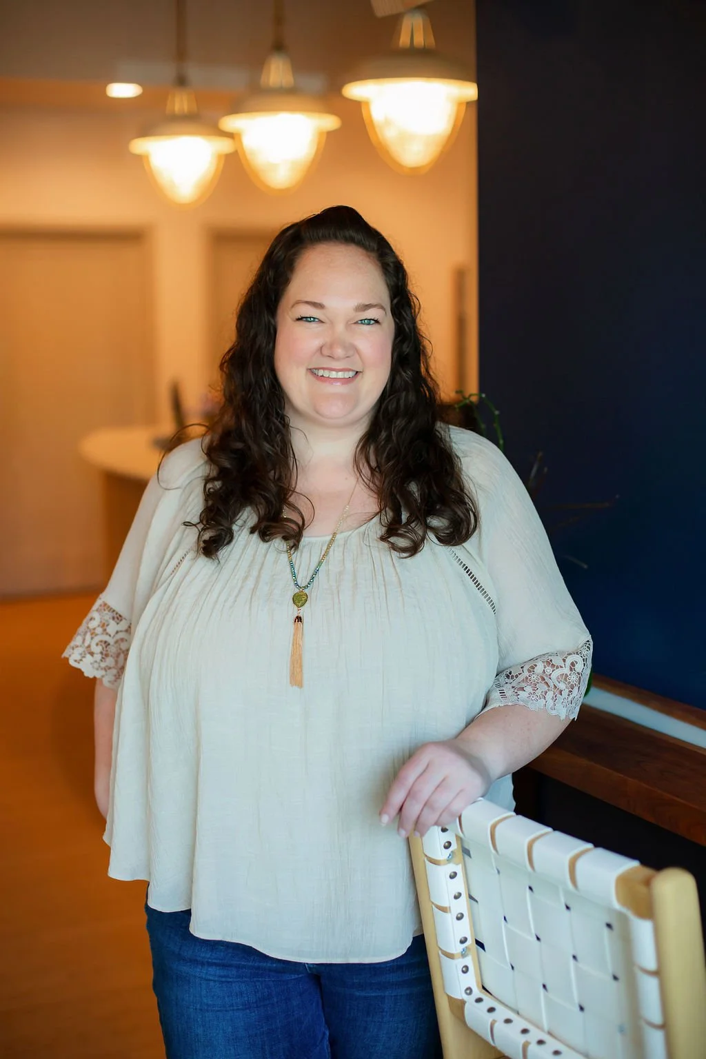 A smiling woman with long, curly dark hair standing indoors near a changing table, with warm lighting and a dark accent wall behind her.